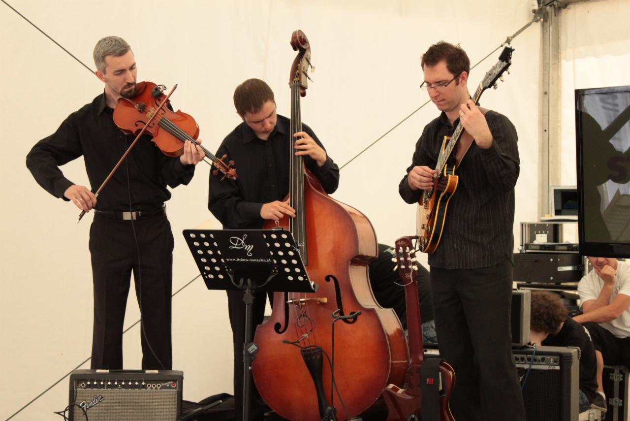 Zespół muzyczny w czarnych strojach grający na skrzypcach, kontrabasie i gitarze elektrycznej na scenie z białym tłem i wzmacniaczami Fender.