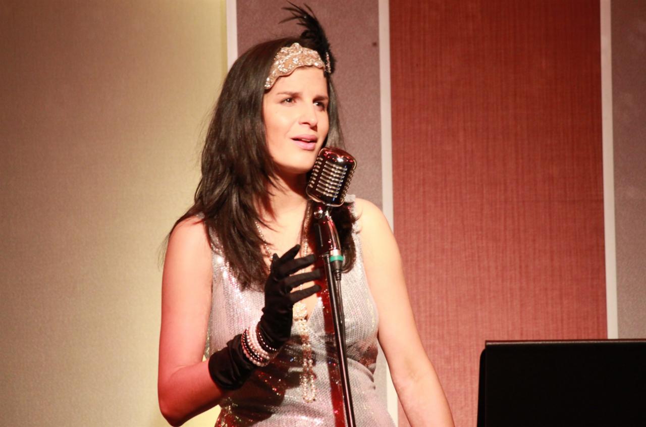 Wokalistka w srebrnej sukience i czarnych rękawiczkach śpiewa do mikrofonu w stylu retro, z ozdobą na głowie z piórem, podczas występu na scenie.