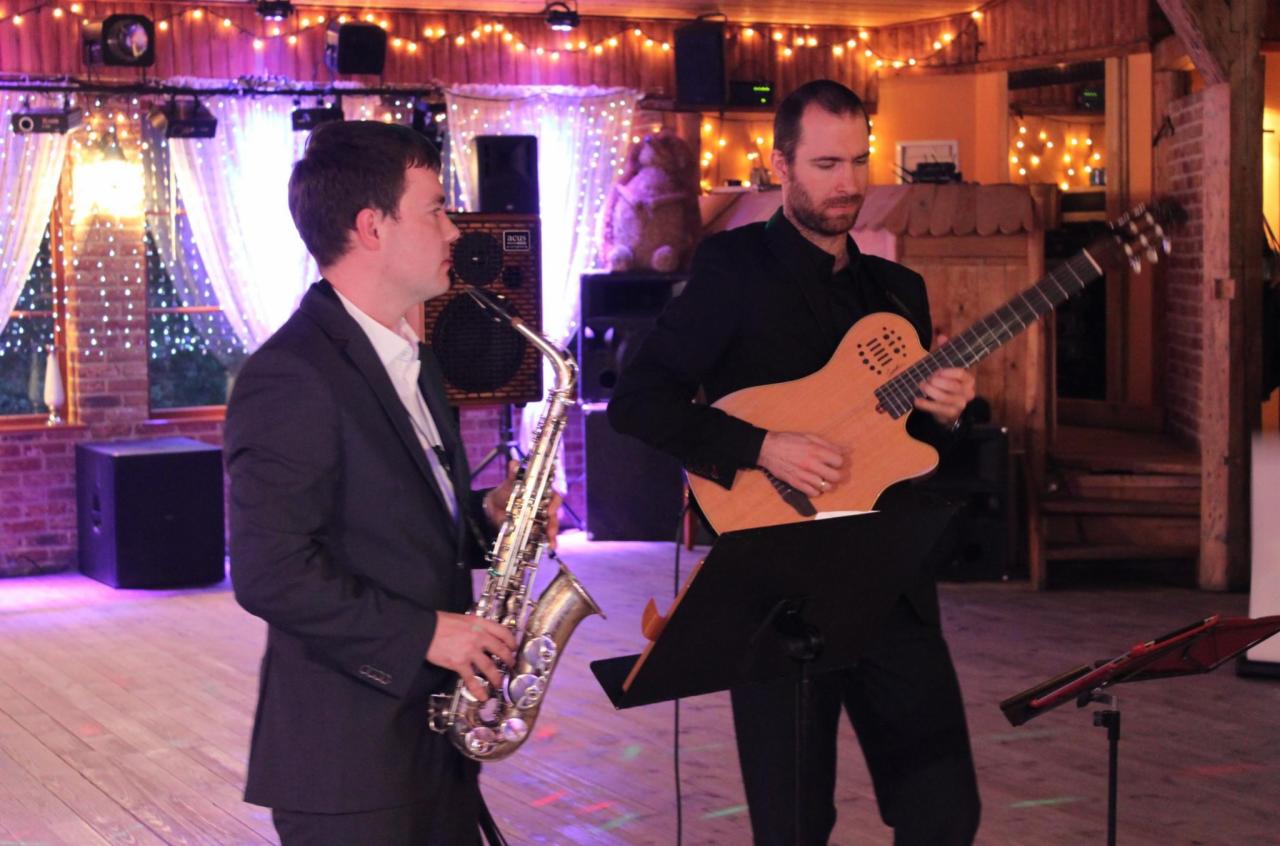 Duet muzyczny: saksofonista i gitarzysta grający na scenie z drewnianą podłogą i oświetleniem dekoracyjnym