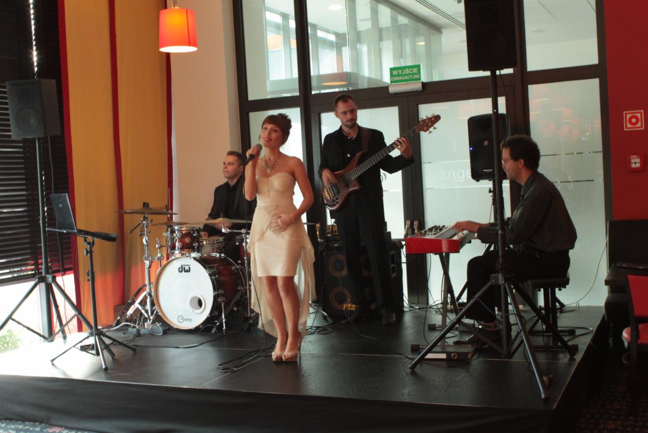 Występ zespołu muzycznego z wokalistką w Katowicach na scenie podczas wydarzenia, widoczny perkusista, basista i klawiszowiec.