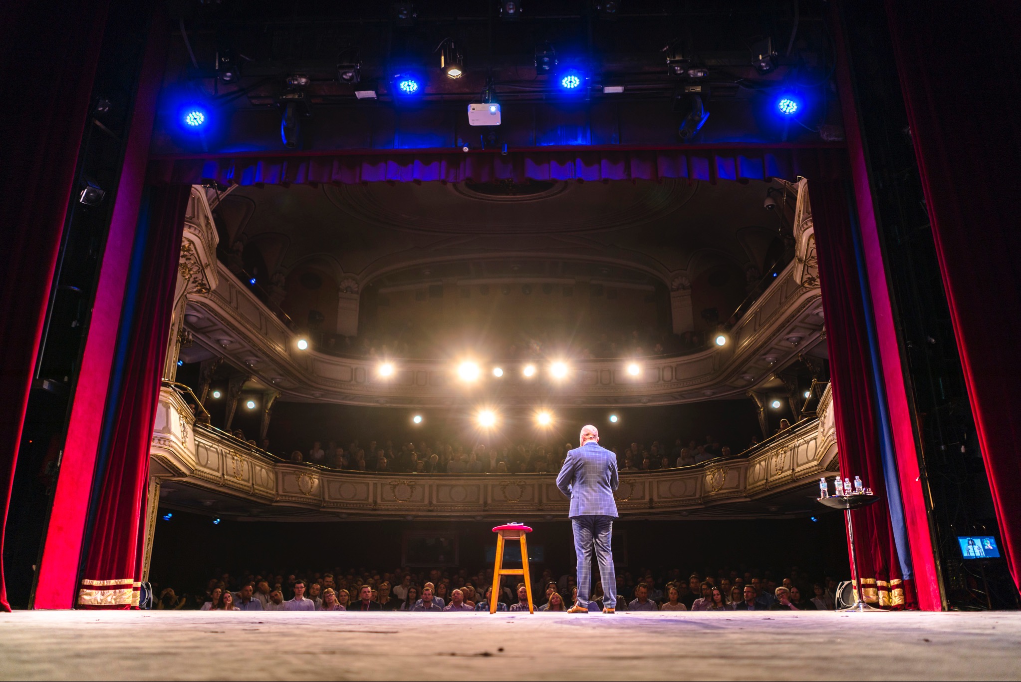 Prelegent w garniturze stoi na scenie przed publicznością w teatrze, z krzesłem i stolikiem z wodą obok, oświetlony reflektorami.