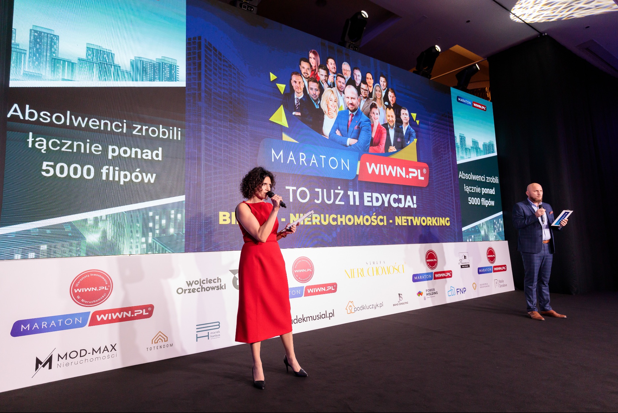 Prelegentka w czerwonej sukience przemawia na scenie podczas konferencji o nieruchomościach, na telebimie wyświetlają się zdjęcia uczestników i informacje o wydarzeniu, w tle widoczny drugi prelegent.