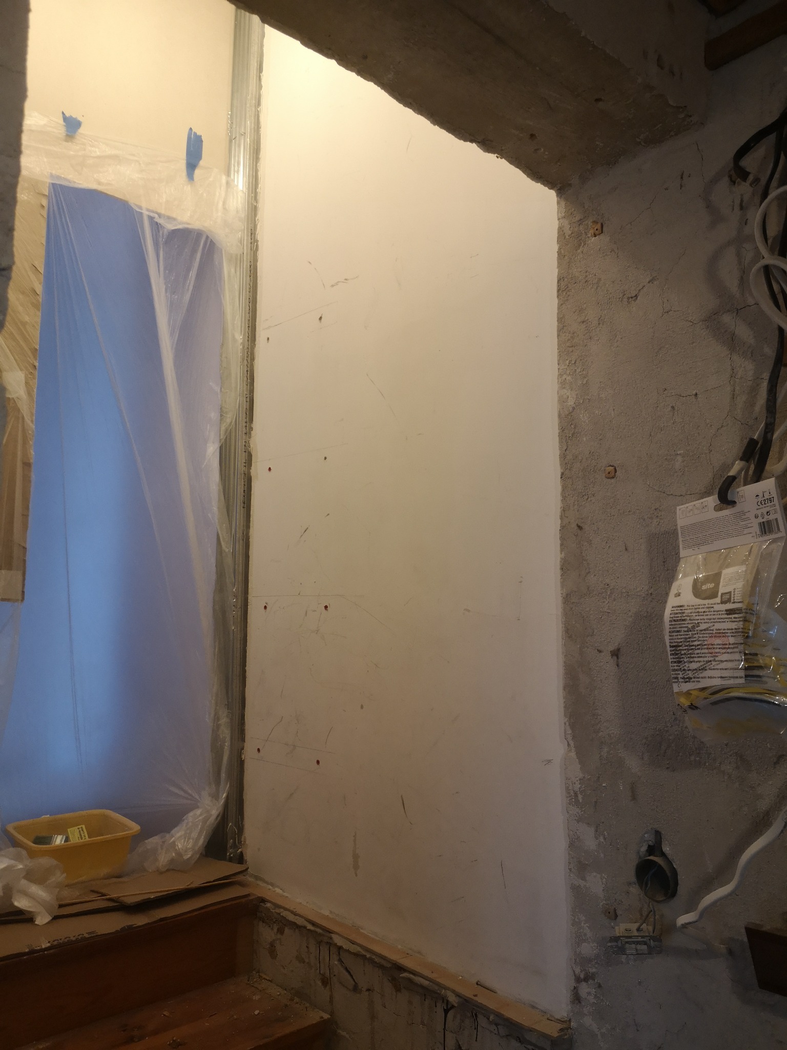 Przygotowanie do gładzi: zabezpieczone folią drzwi, pomalowana ściana z widocznymi otworami po kołkach, surowy betonowy narożnik z wystającymi kablami.