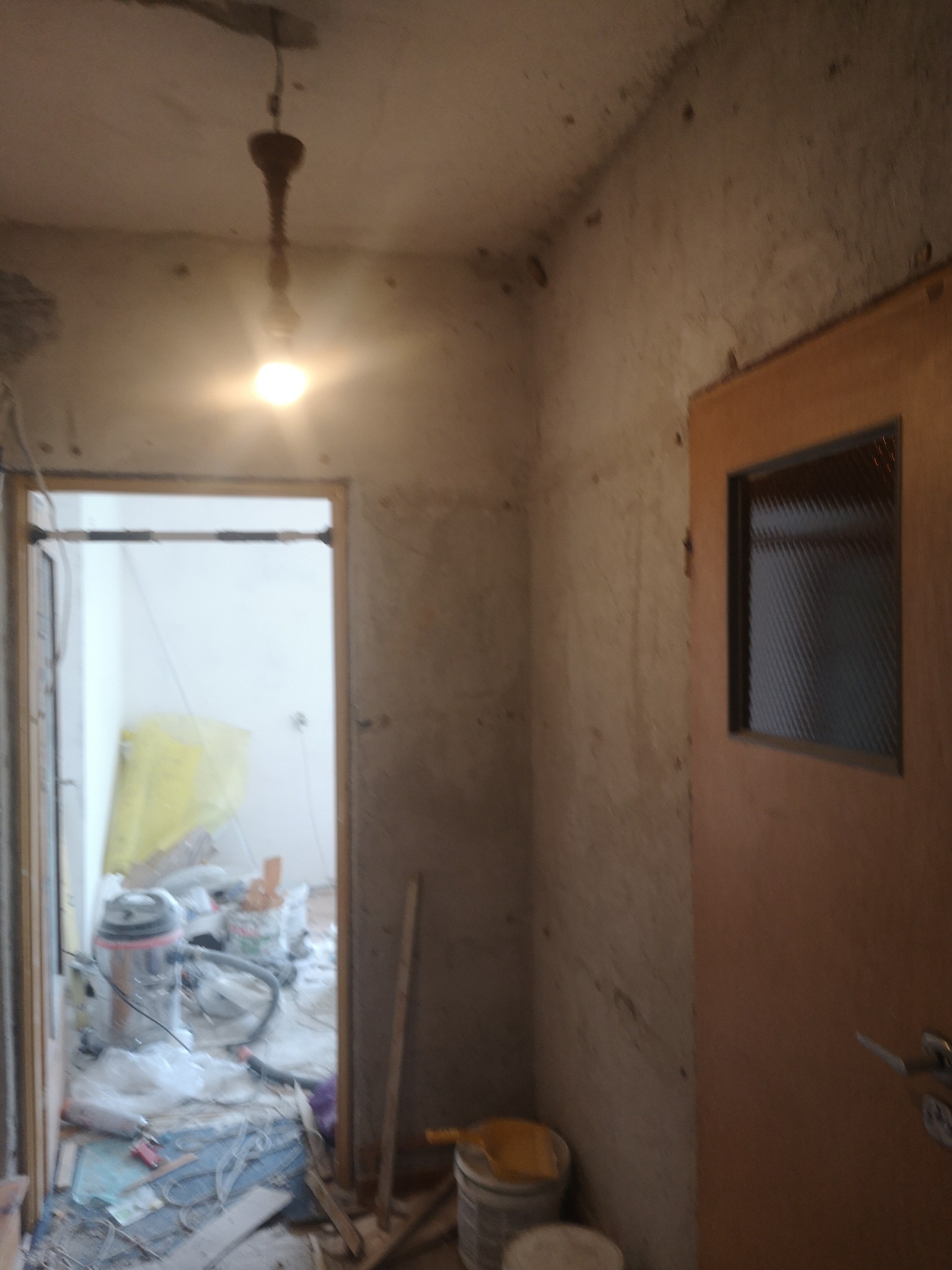Korytarz w trakcie remontu: ściany przed gładzią, widoczny odkurzacz przemysłowy, resztki materiałów budowlanych i stara lampa na suficie.