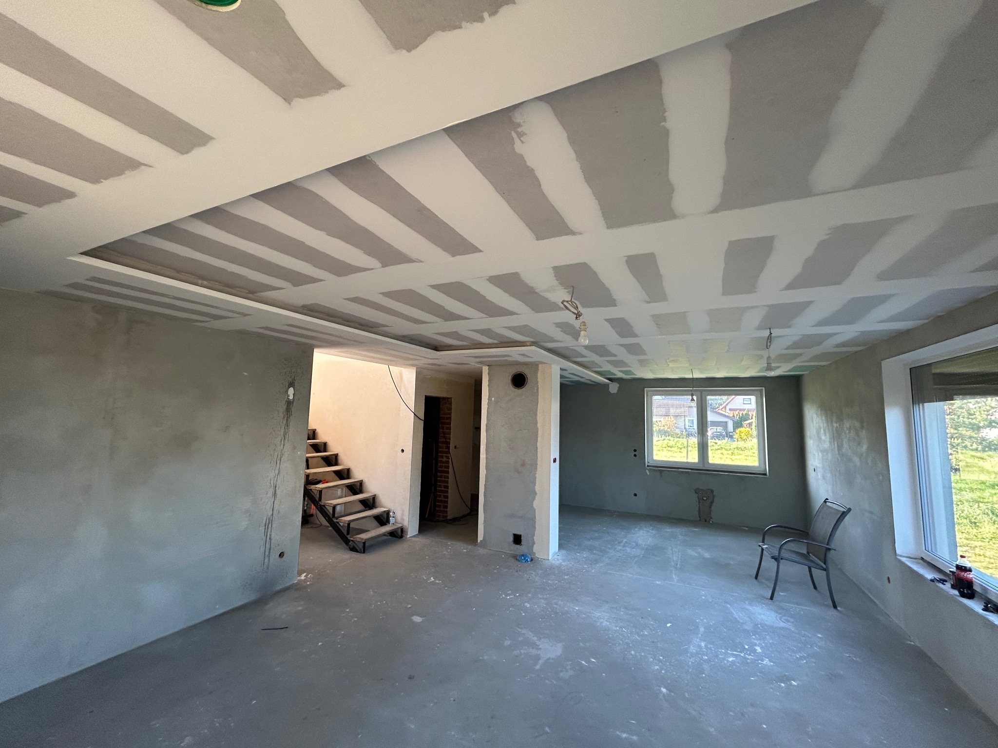 Wnętrze domu w trakcie remontu: surowe ściany z gładzią, odkryte schody, sufit z płyt karton-gips z widocznymi łączeniami, pojedyncze krzesło przy oknie.