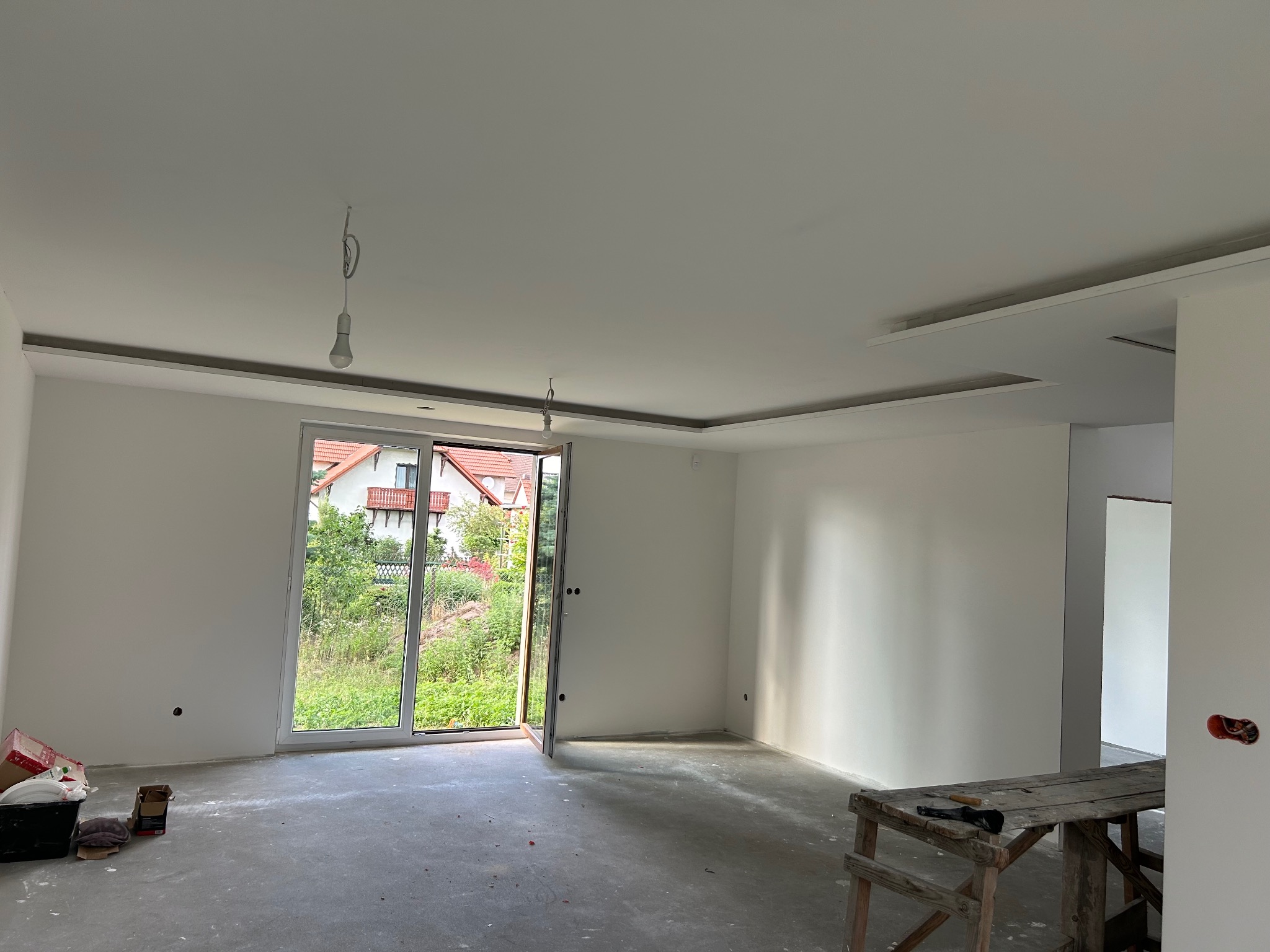 Wnętrze świeżo wykończonego pokoju z pomalowanymi na biało ścianami, widoczne narzędzia malarskie i drabina, z otwartym balkonem z widokiem na zieleń.