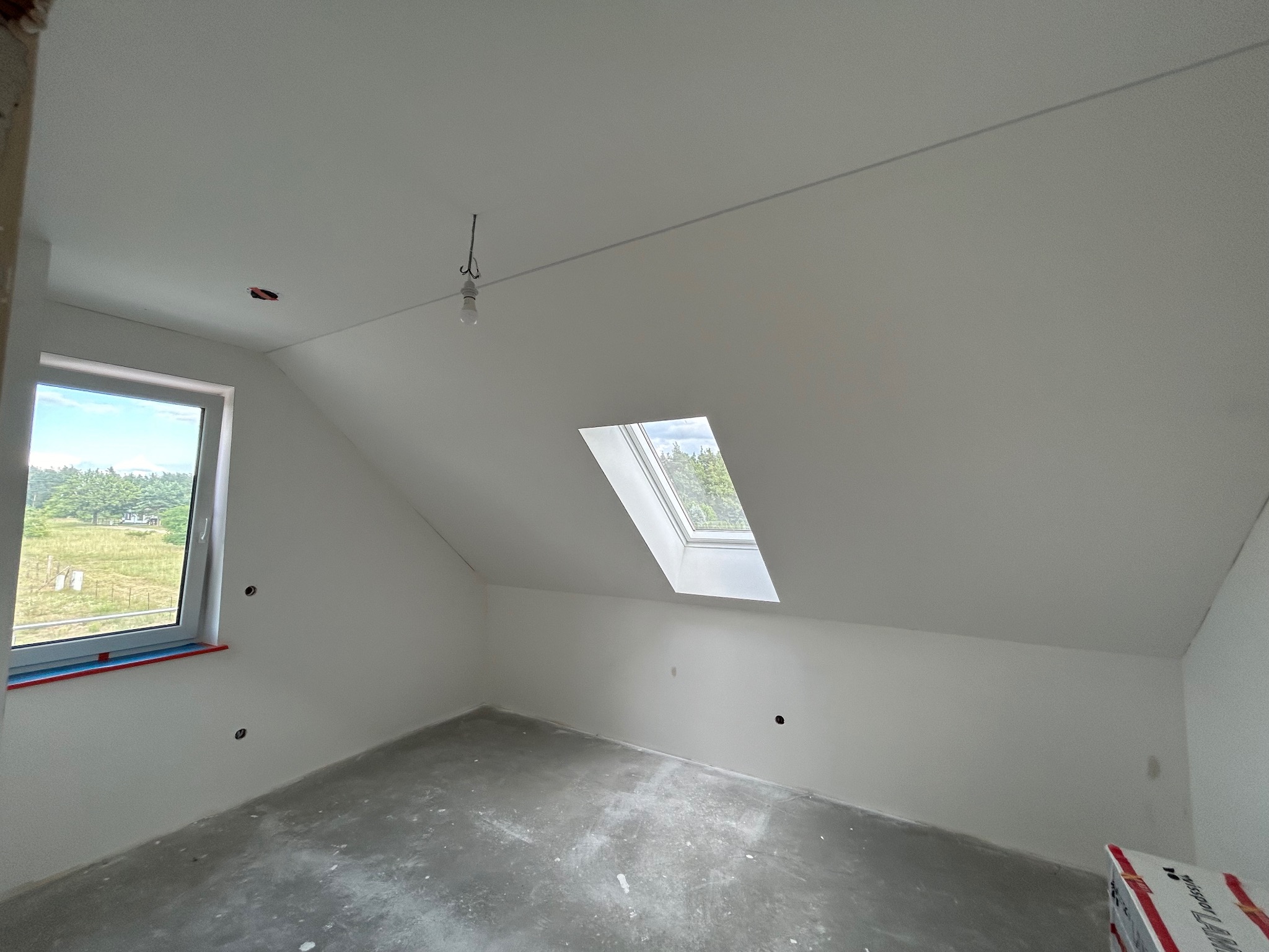 Poddasze w trakcie prac wykończeniowych: świeżo zagruntowane ściany i sufit na biało, surowa betonowa podłoga, okno dachowe wpuszczające światło dzienne, widoczne punkty instalacji elektrycznej...