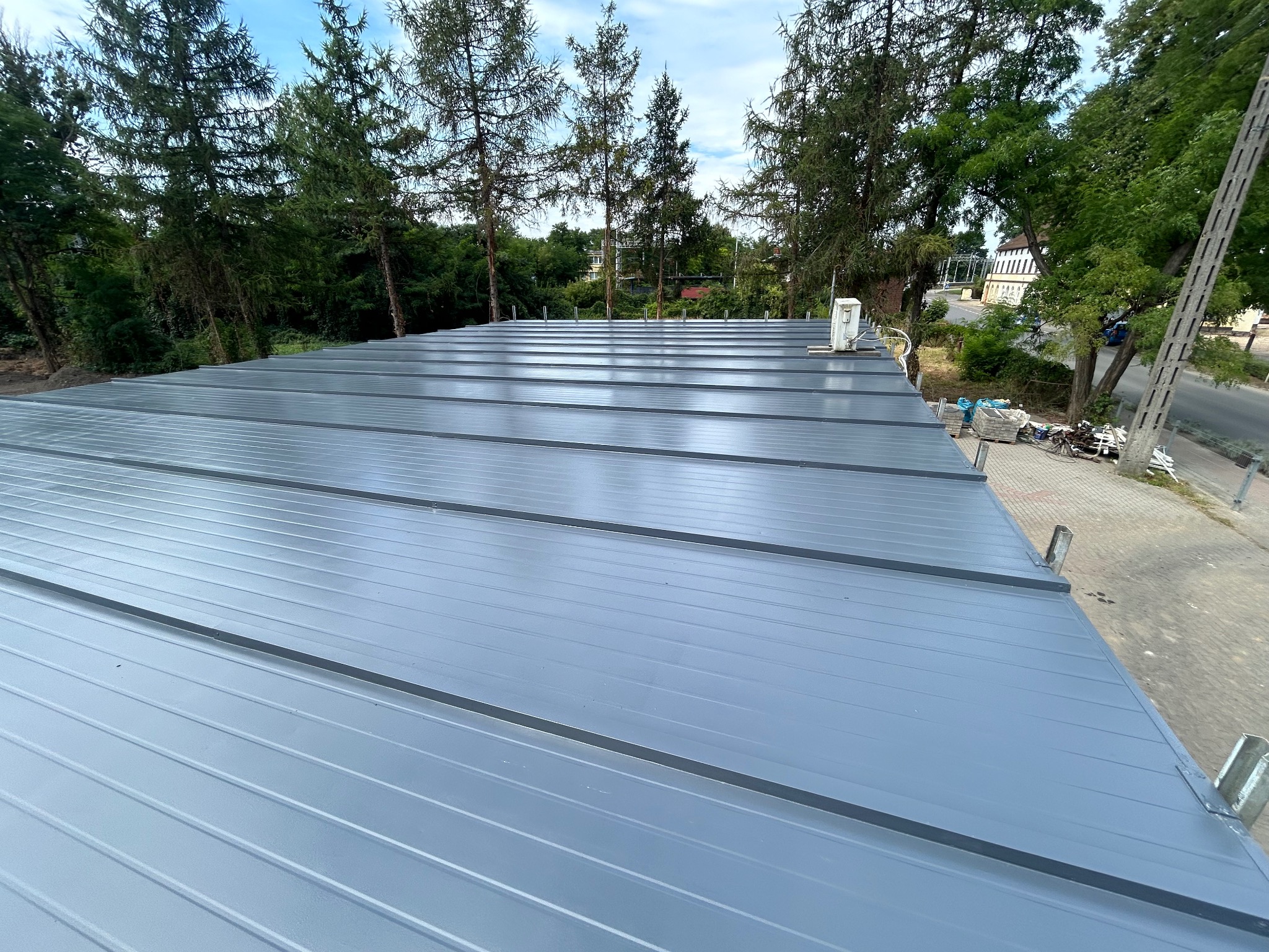 Świeżo pomalowany, blaszany dach w odcieniach szarości z widocznym systemem rynnowym, otoczony zielenią drzew i elementami infrastruktury miejskiej.
