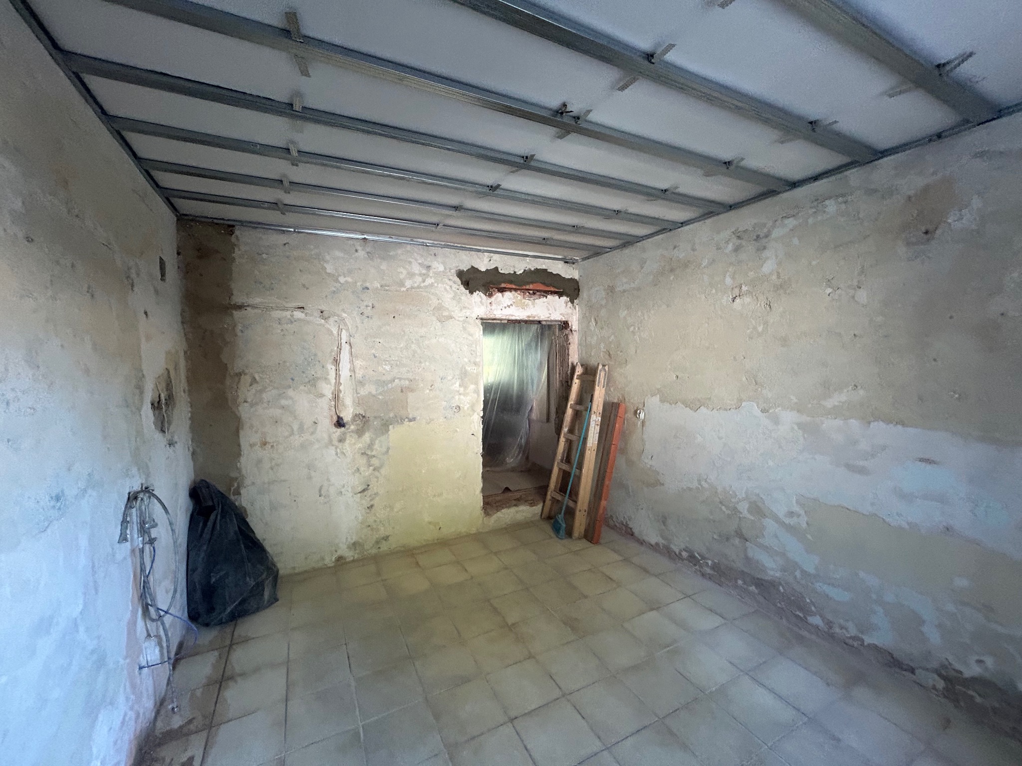 Pokój w trakcie remontu: widoczne stare ściany, metalowa konstrukcja sufitu podwieszanego z płyt karton-gips, częściowo przygotowane do montażu, oraz drabina oparta o futrynę zabezpieczoną folią.