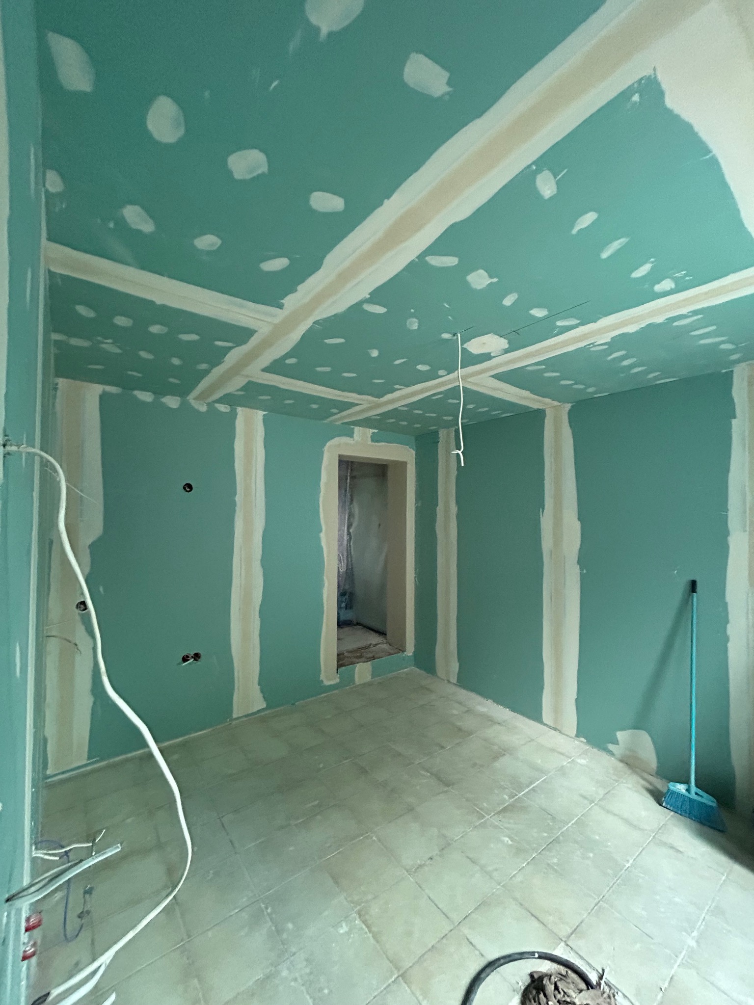 Pokój w trakcie prac wykończeniowych: ściany i sufit pokryte zieloną farbą, z widocznymi białymi paskami wzmacniającymi, miejscami zaszpachlowane otwory, na podłodze widoczne płytki, a w rogu stoi...