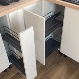 Firma Alppa - Montaż wysuwanych koszy cargo w szafce kuchennej, widoczne narzędzia i akcesoria na blacie, podłoga imitująca drewno.