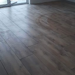 Firma Alppa - Ułożona podłoga z paneli imitujących drewno w surowym pomieszczeniu z widocznymi śladami po okręgu, okno balkonowe z widokiem na zaśnieżony teren, wystające kable elektryczne.