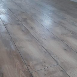 Firma Alppa - Ułożone panele podłogowe w jasnym odcieniu drewna, widoczne fugi i fragment ściany z gniazdkami elektrycznymi.