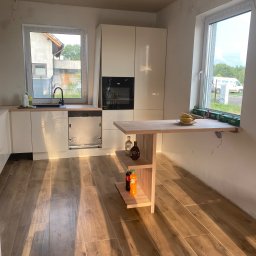 Firma Alppa - Wnętrze kuchni w trakcie prac wykończeniowych, widoczna zabudowa kuchenna z drewnianym blatem, drewniana podłoga, stół przymocowany do ściany, a na nim banany i puszki, ściany częściowo wykończone,...