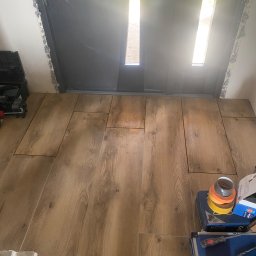 Firma Alppa - Układanie podłogi z paneli drewnopodobnych przy wejściu, widoczne narzędzia i taśmy malarskie, drzwi wejściowe z widocznym światłem dziennym.