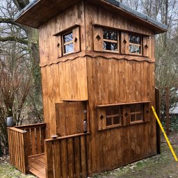 Firma Alppa - Drewniana konstrukcja przypominająca domek na drzewie z małym balkonem i oknami z ozdobnymi wykończeniami, widoczny poziomica po prawej stronie, butelka na balkonie.