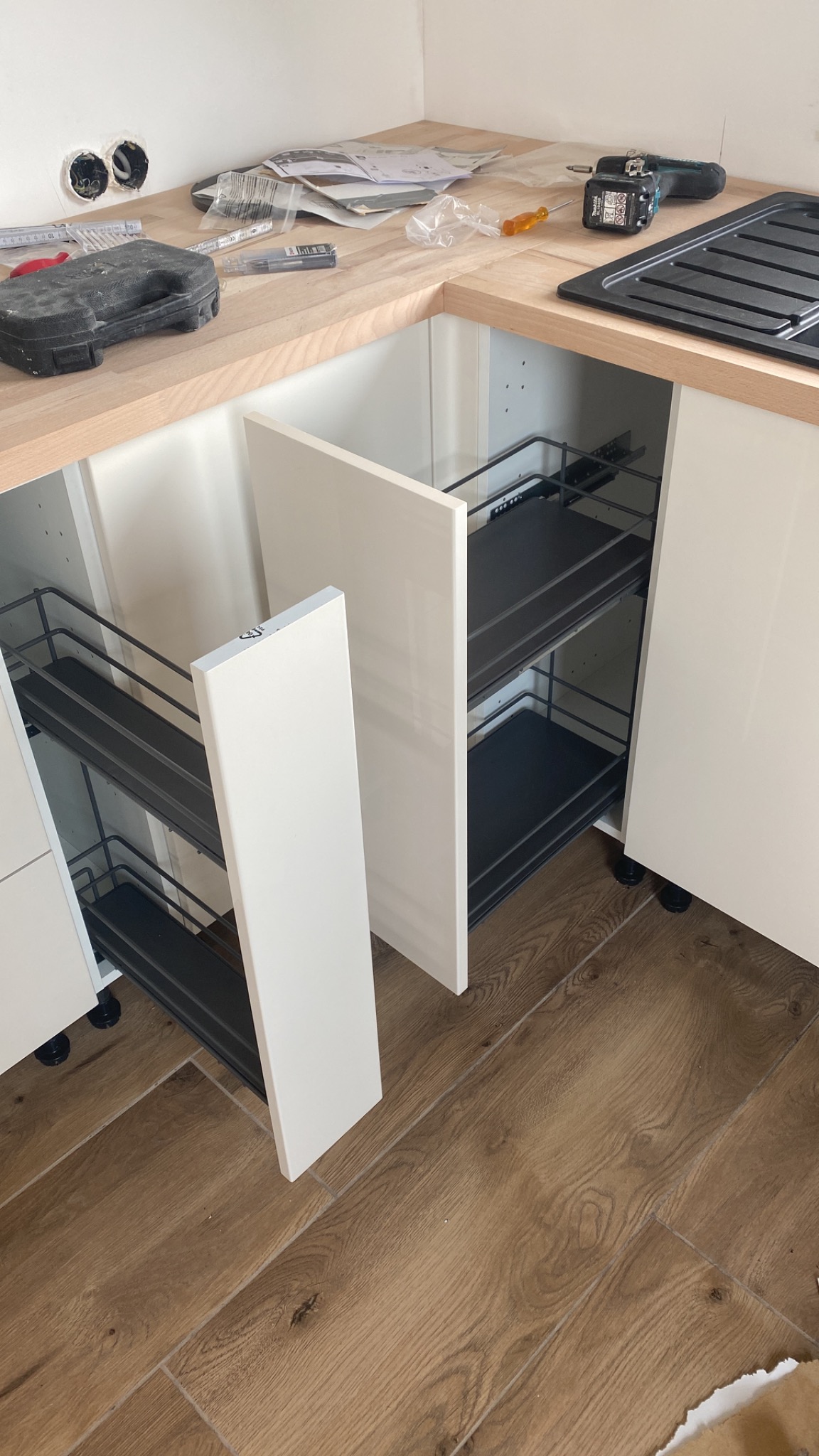 Montaż wysuwanych koszy cargo w szafce kuchennej, widoczne narzędzia i akcesoria na blacie, podłoga imitująca drewno.