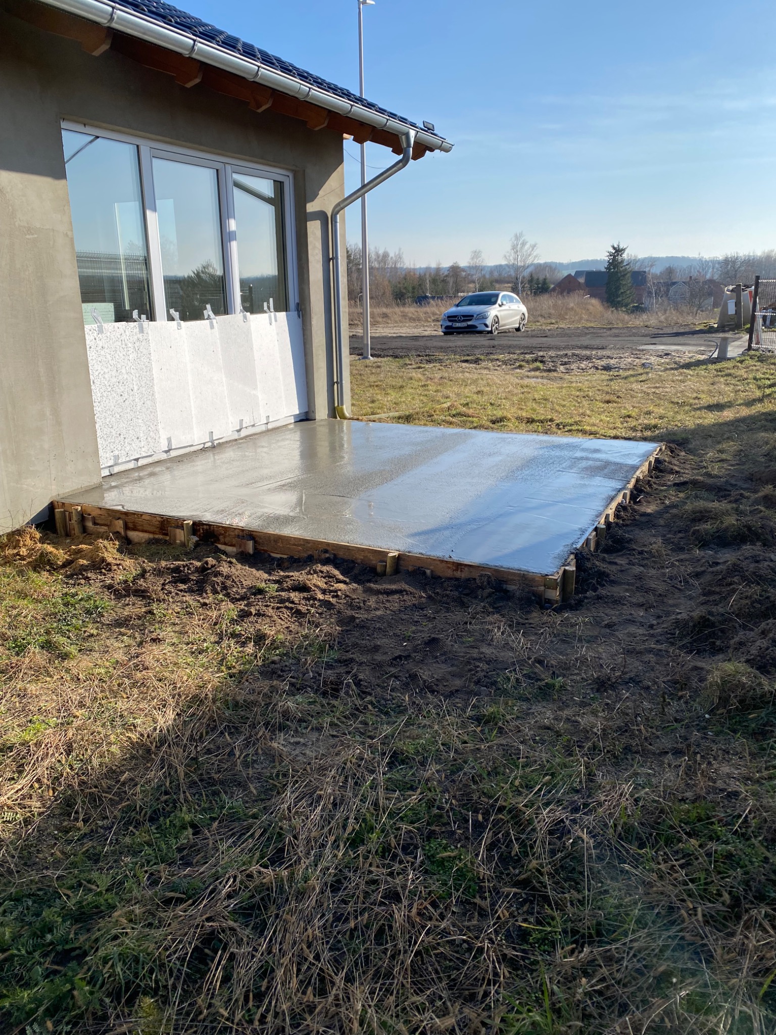 Wylana, świeża betonowa wylewka pod taras, otoczona drewnianym szalunkiem, przylegająca do ściany budynku z oknem i fragmentami styropianu, w tle samochód osobowy.