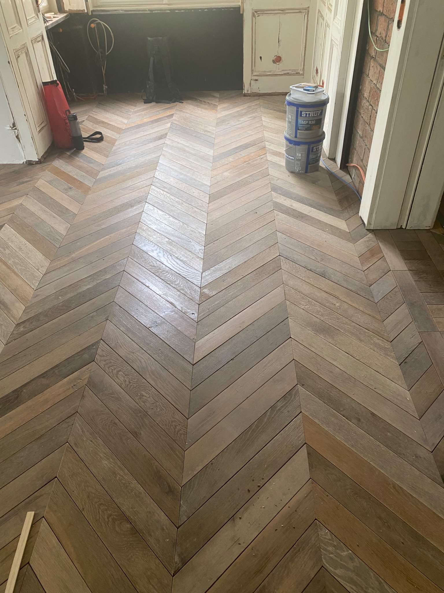 Ułożony parkiet w jodełkę, widoczne wiaderka z klejem Stauf, elementy wyposażenia w tle.