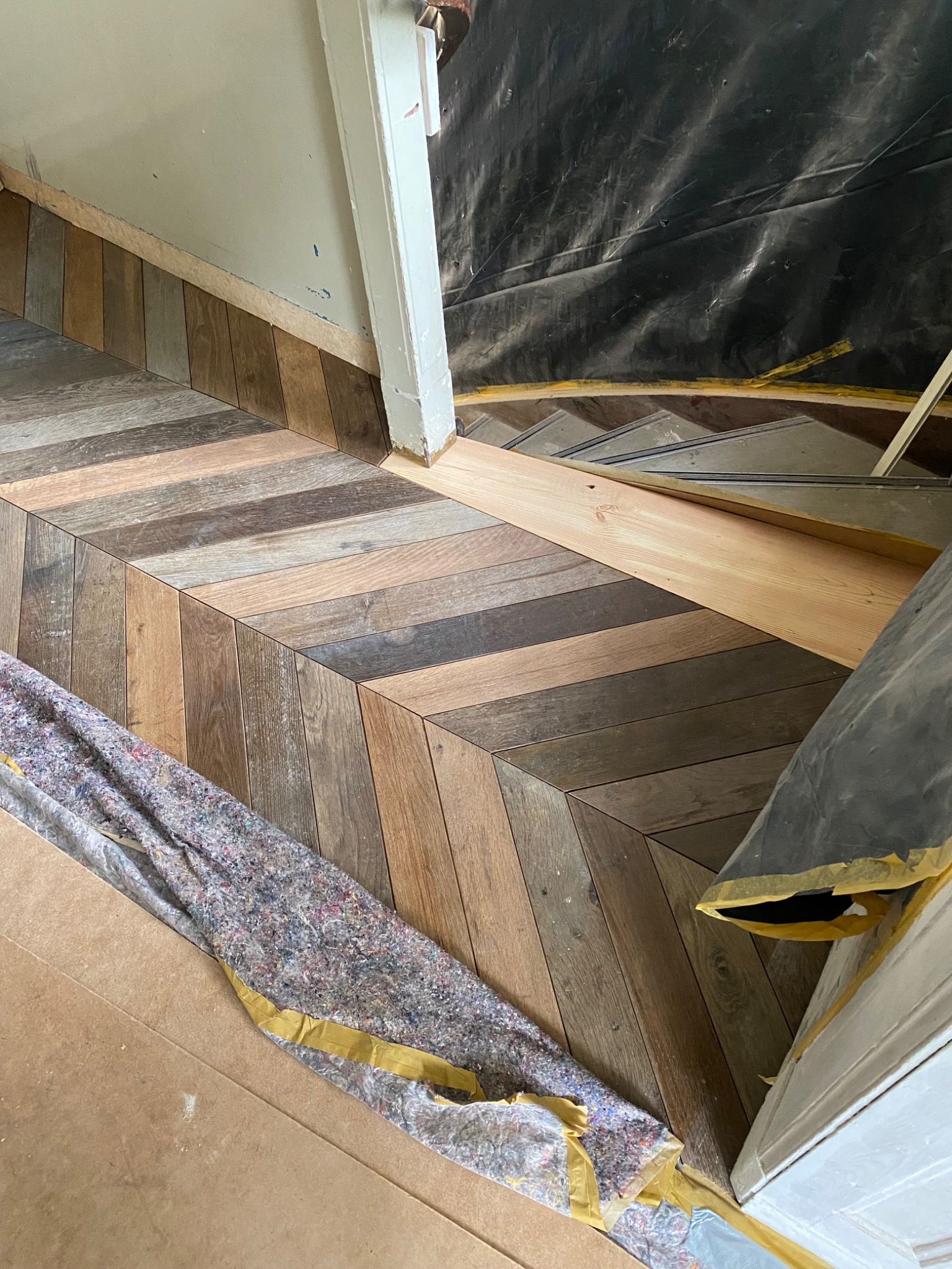 Fragment podłogi z ułożonym parkietem w jodełkę w odcieniach brązu, zabezpieczona folią i materiałem ochronnym podczas prac wykończeniowych przy ścianie i framudze drzwi.