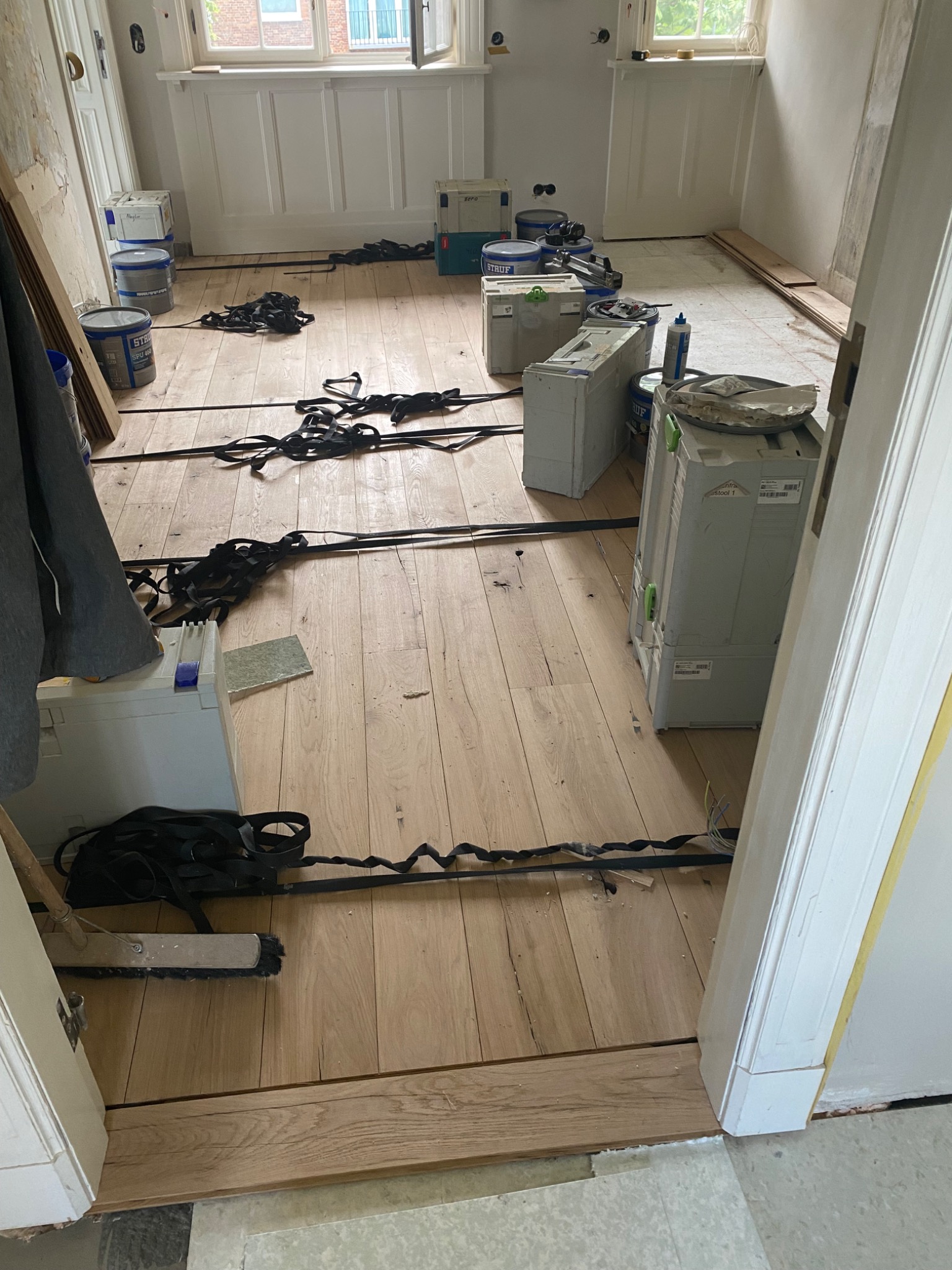 Wnętrze w trakcie układania drewnianej podłogi, widoczne narzędzia, puszki z klejem, skrzynki narzędziowe i czarne pasy podtrzymujące klejone deski.