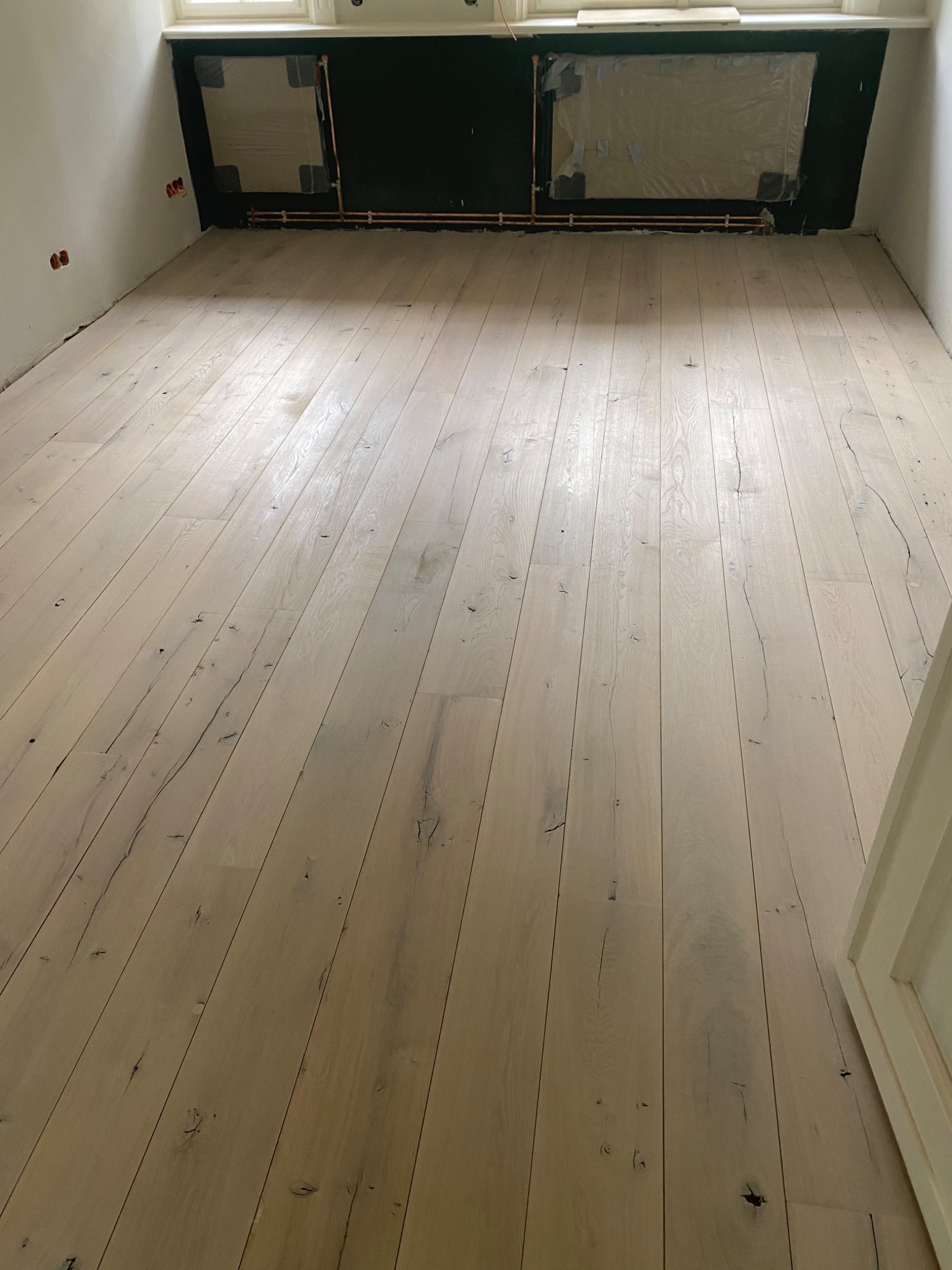 Jasna, drewniana podłoga z widocznymi sękami i pęknięciami, ułożona w pustym pokoju z przygotowanymi otworami okiennymi i instalacją grzewczą.