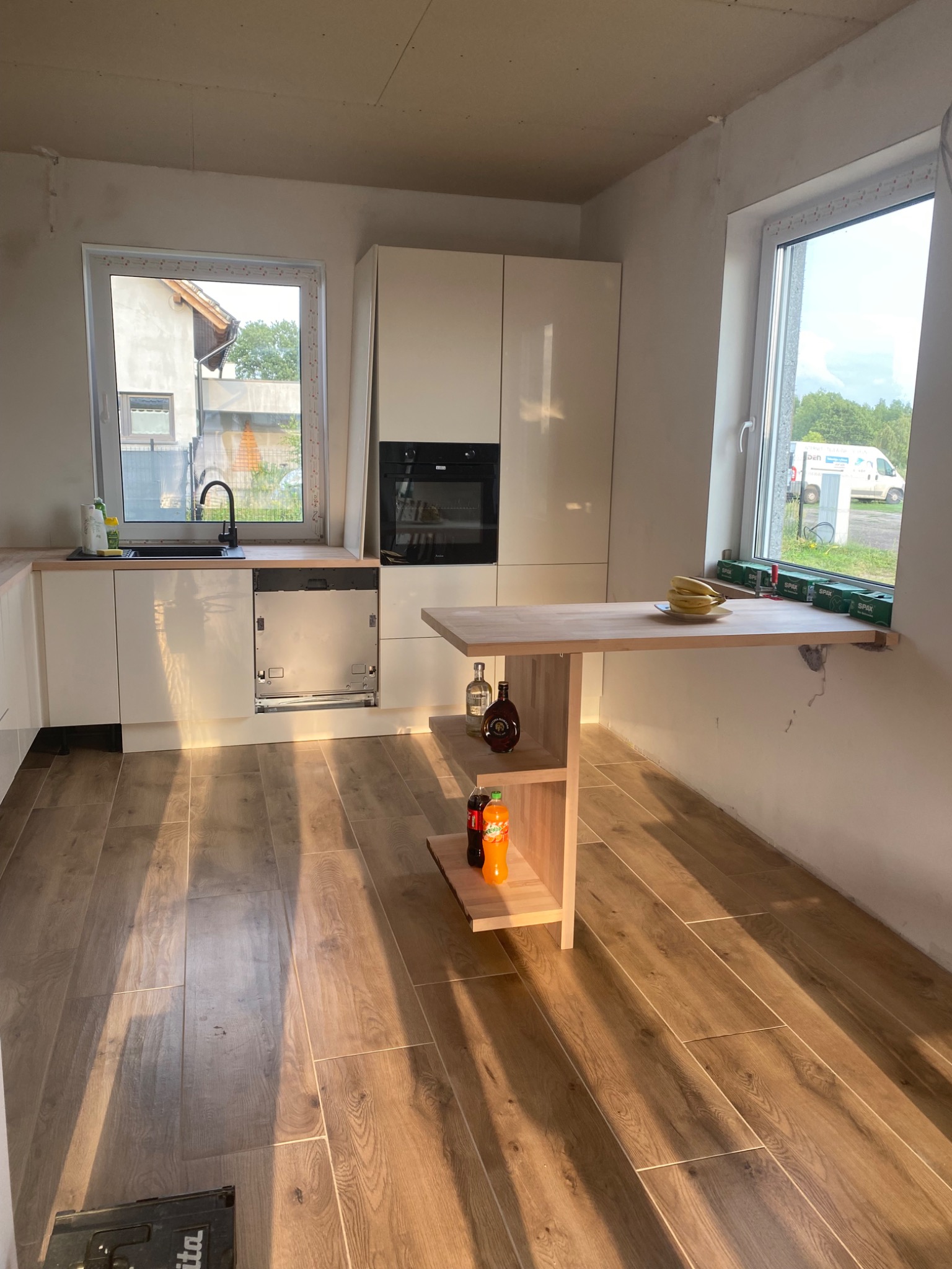 Wnętrze kuchni w trakcie prac wykończeniowych, widoczna zabudowa kuchenna z drewnianym blatem, drewniana podłoga, stół przymocowany do ściany, a na nim banany i puszki, ściany częściowo wykończone,...