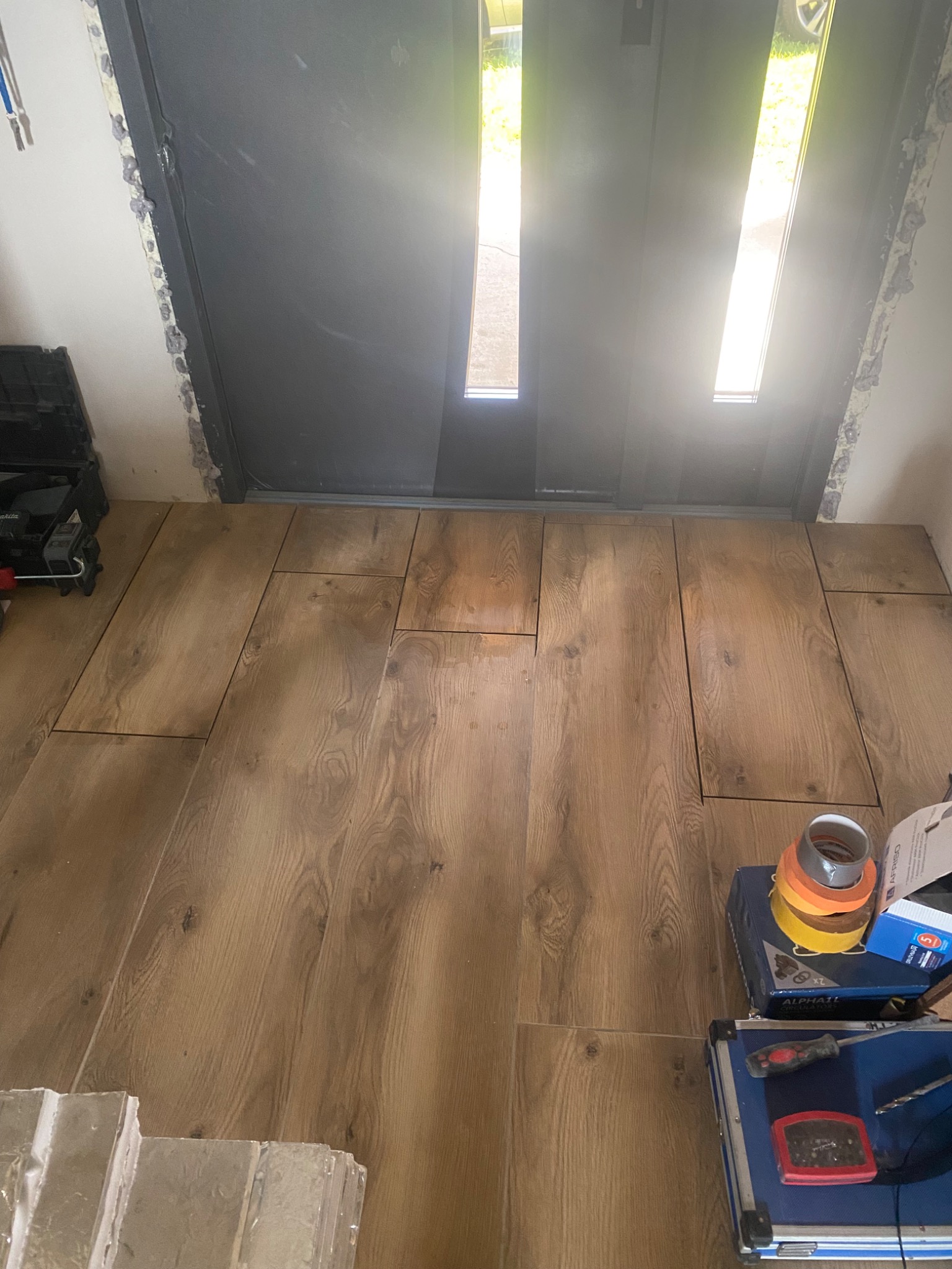 Układanie podłogi z paneli drewnopodobnych przy wejściu, widoczne narzędzia i taśmy malarskie, drzwi wejściowe z widocznym światłem dziennym.