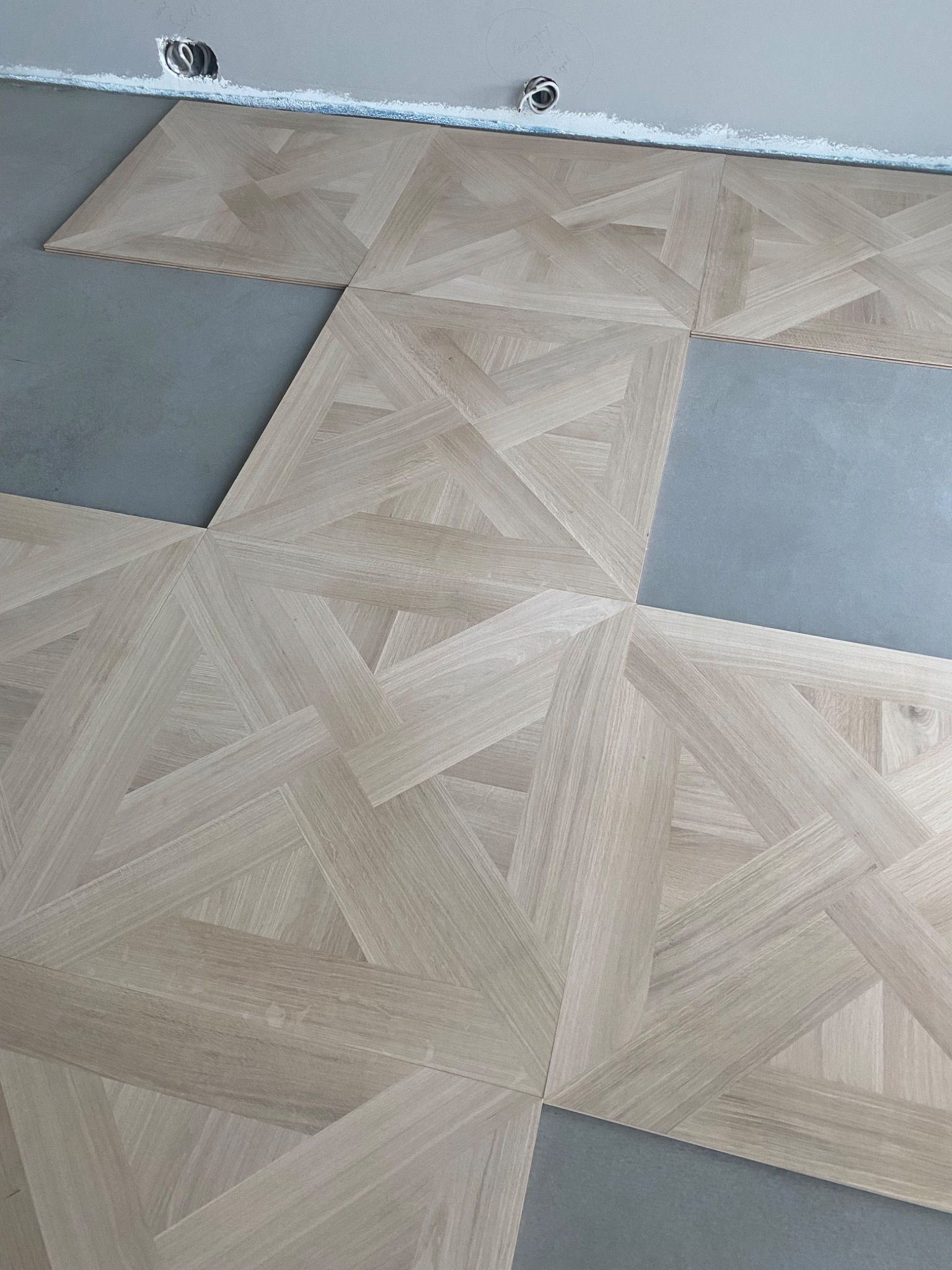 Fragment układania parkietu w geometryczny wzór, widoczne niedokończone fragmenty podłogi i surowy sufit z instalacją elektryczną.