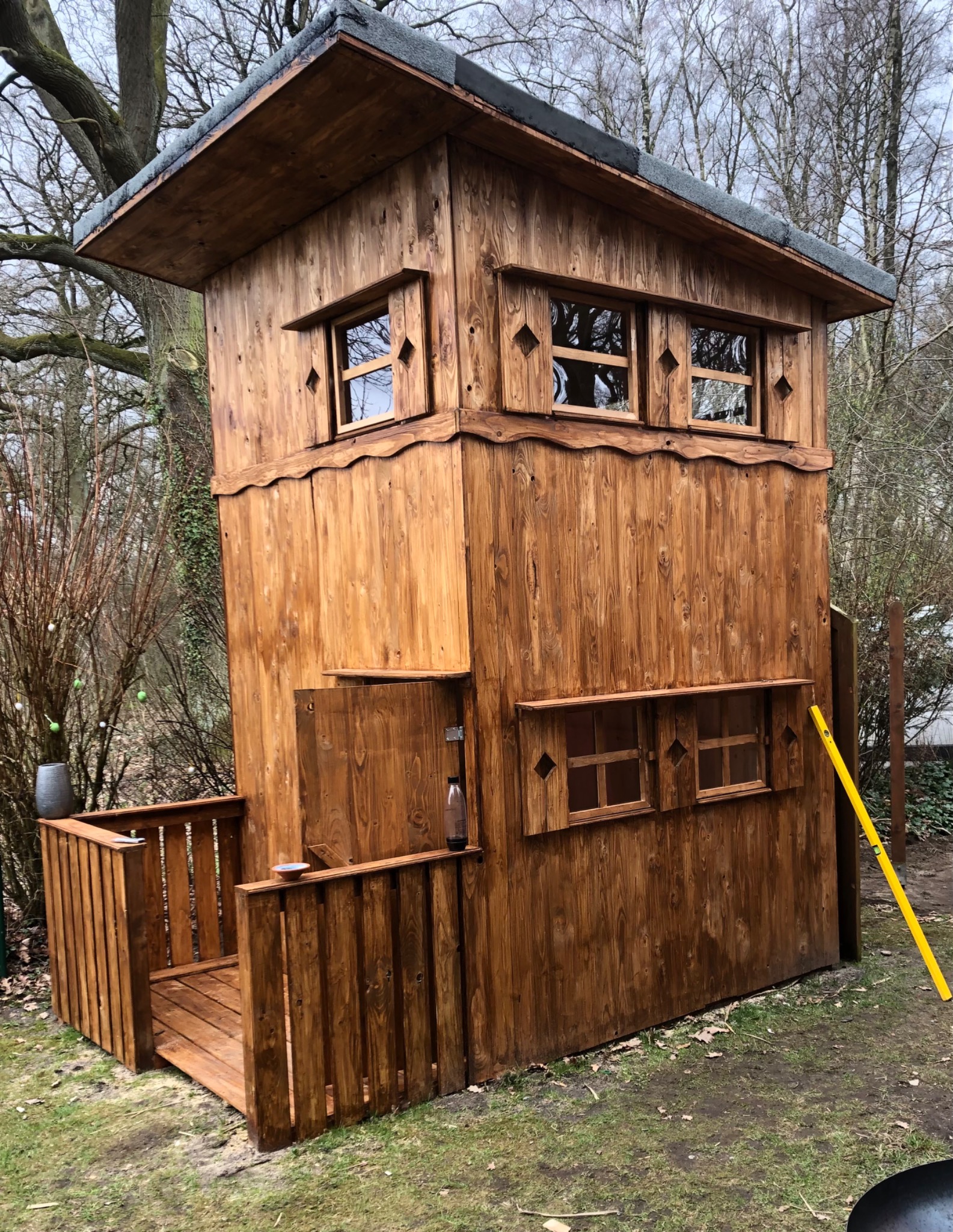 Drewniana konstrukcja przypominająca domek na drzewie z małym balkonem i oknami z ozdobnymi wykończeniami, widoczny poziomica po prawej stronie, butelka na balkonie.