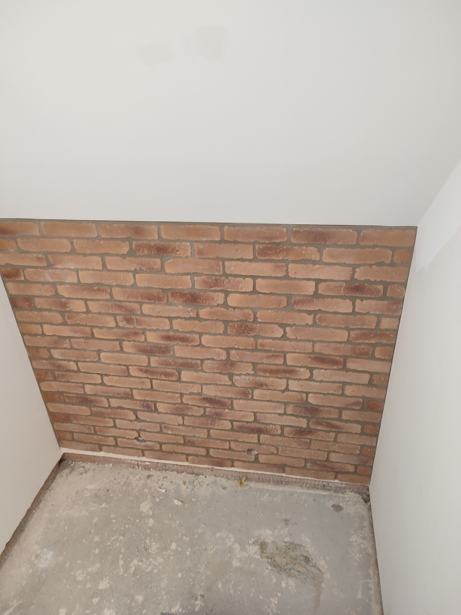 Ściana wykończona cegłą dekoracyjną w trakcie prac, surowy beton na podłodze, białe ściany i sufit, widok z dołu, oświetlenie dzienne.