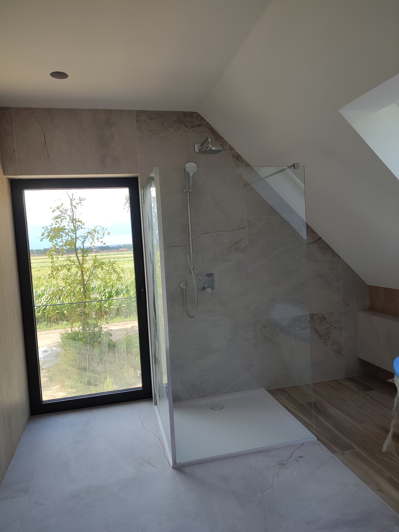 Kabina prysznicowa ze szklaną ścianką w łazience na poddaszu, z oknem z widokiem na pole.