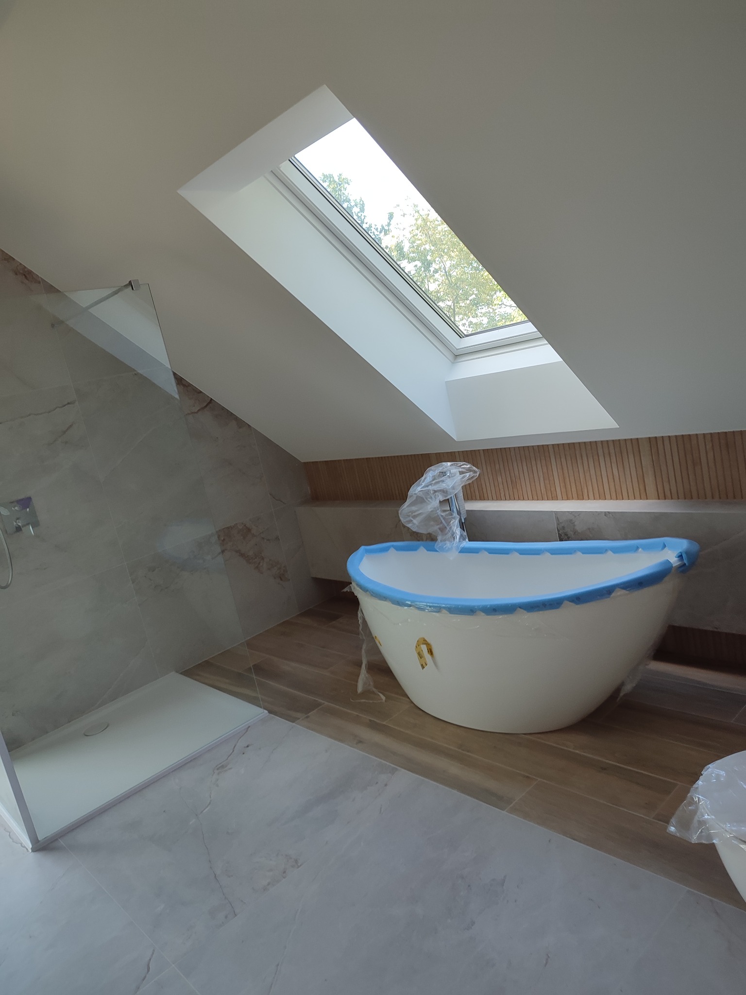 Nowa, owalna wanna zabezpieczona folią i niebieską taśmą malarską w łazience na poddaszu ze skośnym oknem dachowym, obok kabina prysznicowa ze szklaną ścianką.