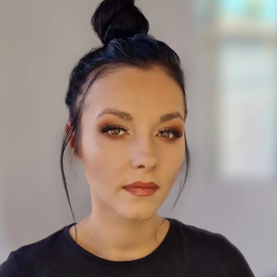 Portret kobiety z profesjonalnie wykonanym makijażem oczu w odcieniach brązu i beżu, z błyszczącymi ustami i włosami upiętymi w kok. Widoczne pojedyncze kosmyki włosów opadające na twarz.