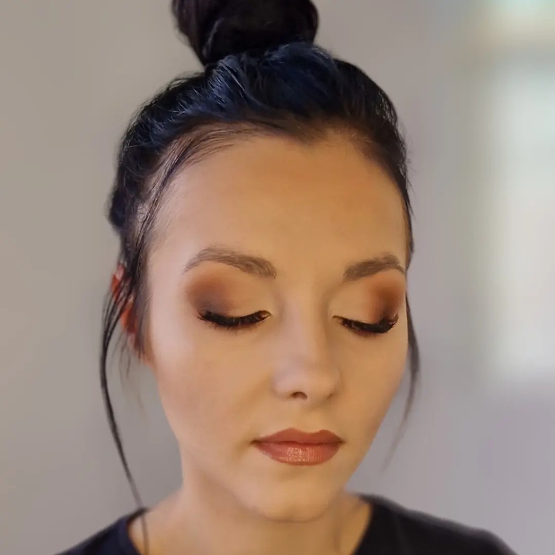 Portret kobiety z subtelnym, brązowym cieniowaniem powiek i błyszczącymi ustami, włosy upięte w wysoki kok.