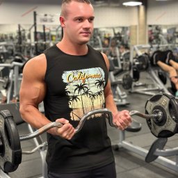 Filip Kręcichwost FK Sport - Mężczyzna w czarnej koszulce z nadrukiem California ćwiczy biceps sztangą łamaną w siłowni. Widoczne maszyny i osoby w tle, oświetlenie sufitowe.