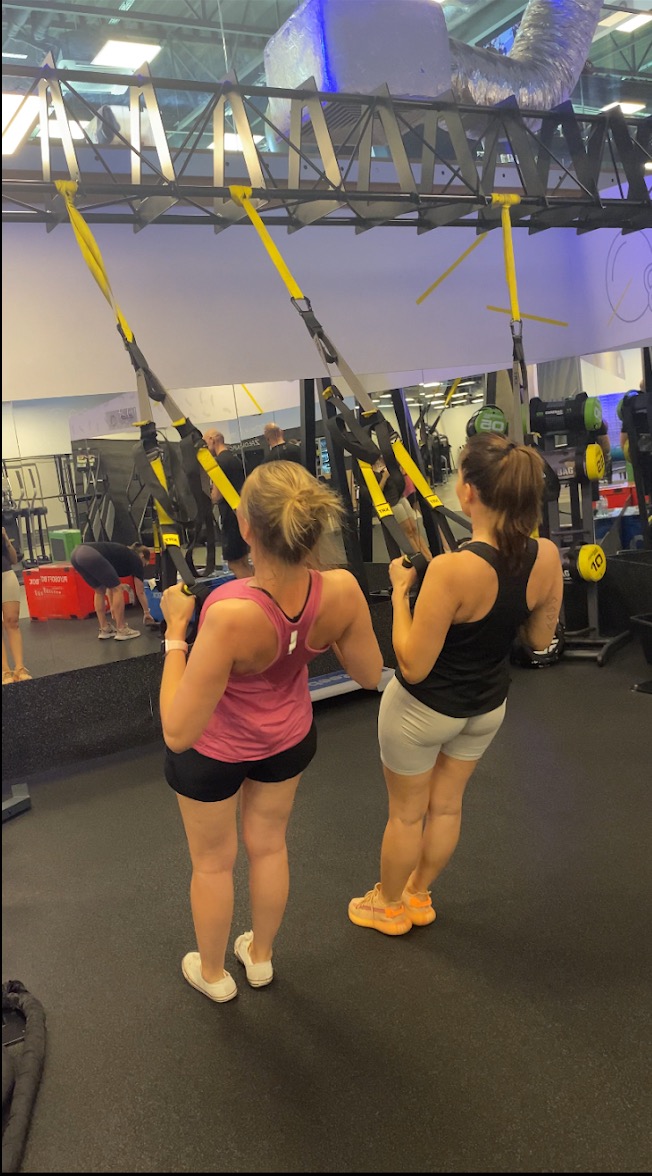 Dwie kobiety ćwiczą na TRX w siłowni, widok z tyłu, jedna w różowej koszulce i czarnych spodenkach, druga w czarnej koszulce i szarych spodenkach, obie trzymają żółte liny TRX.
