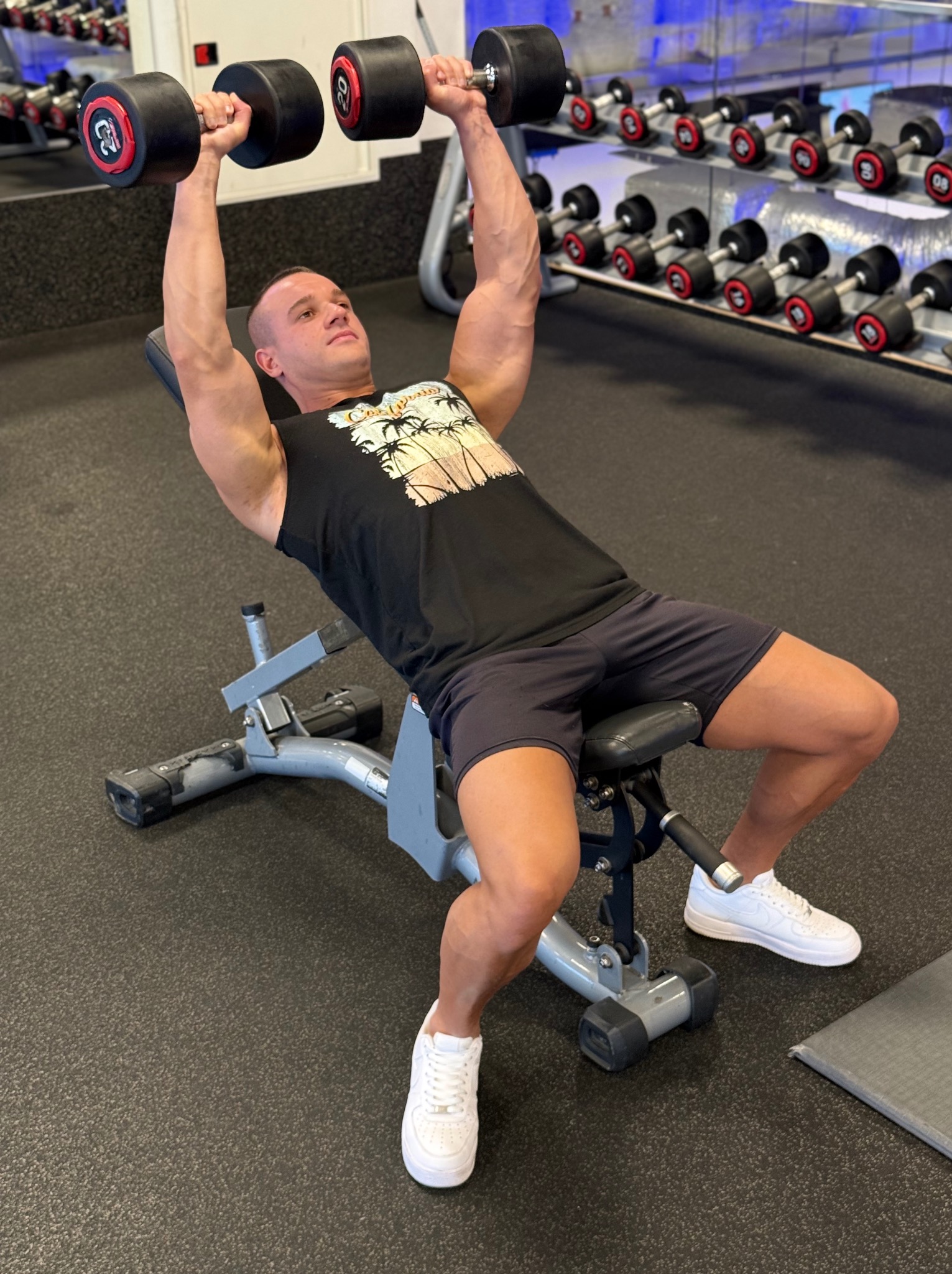 Trener personalny na ławce skośnej, podnosi hantle 25 kg, w tle regał z obciążeniami. Widoczny wysiłek na twarzy, siłownia z lustrami.