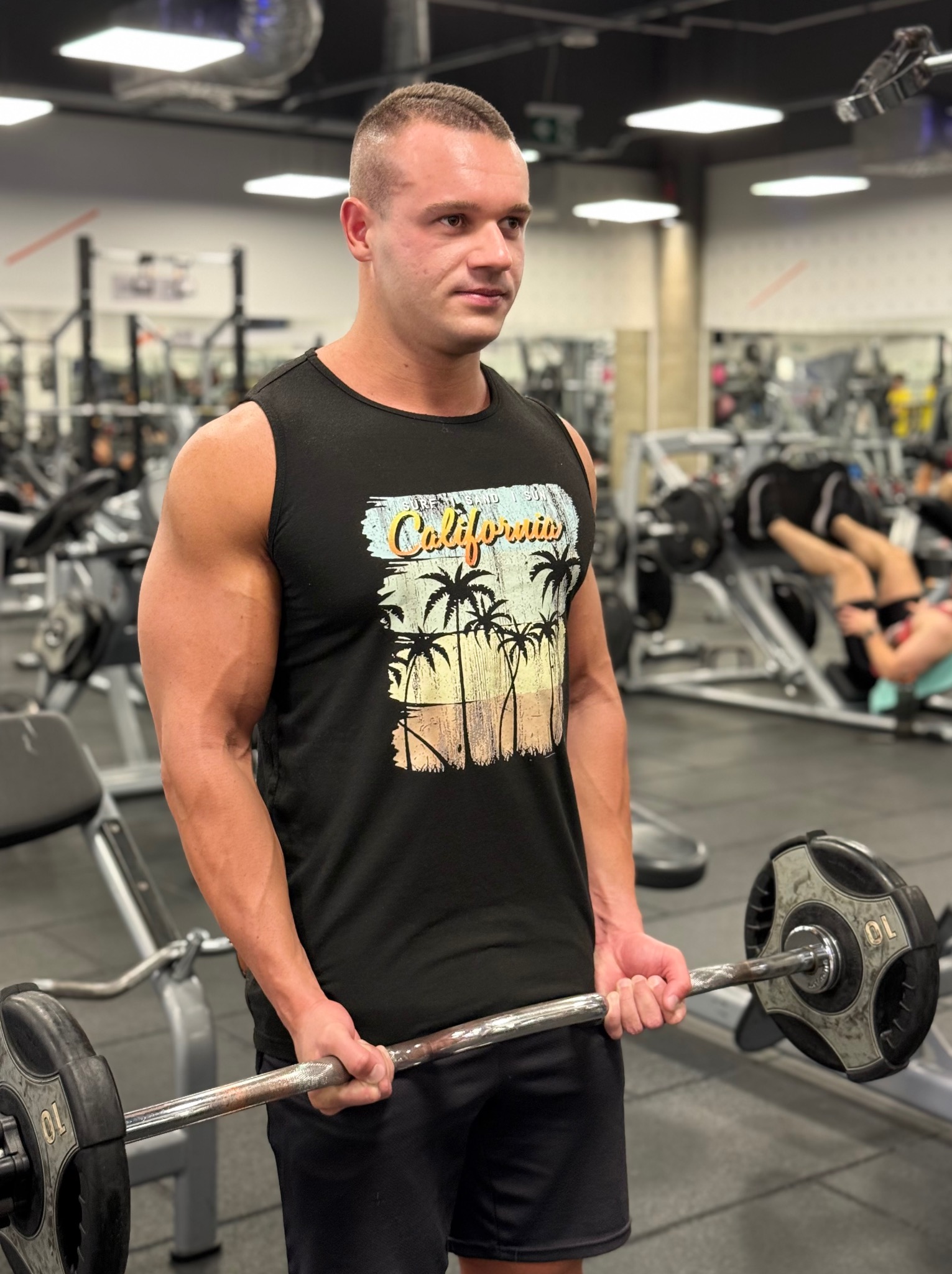 Mężczyzna w czarnym tank topie z nadrukiem 'California' ćwiczy biceps sztangą w siłowni. Widoczne maszyny i osoby w tle. Oświetlenie sufitowe.