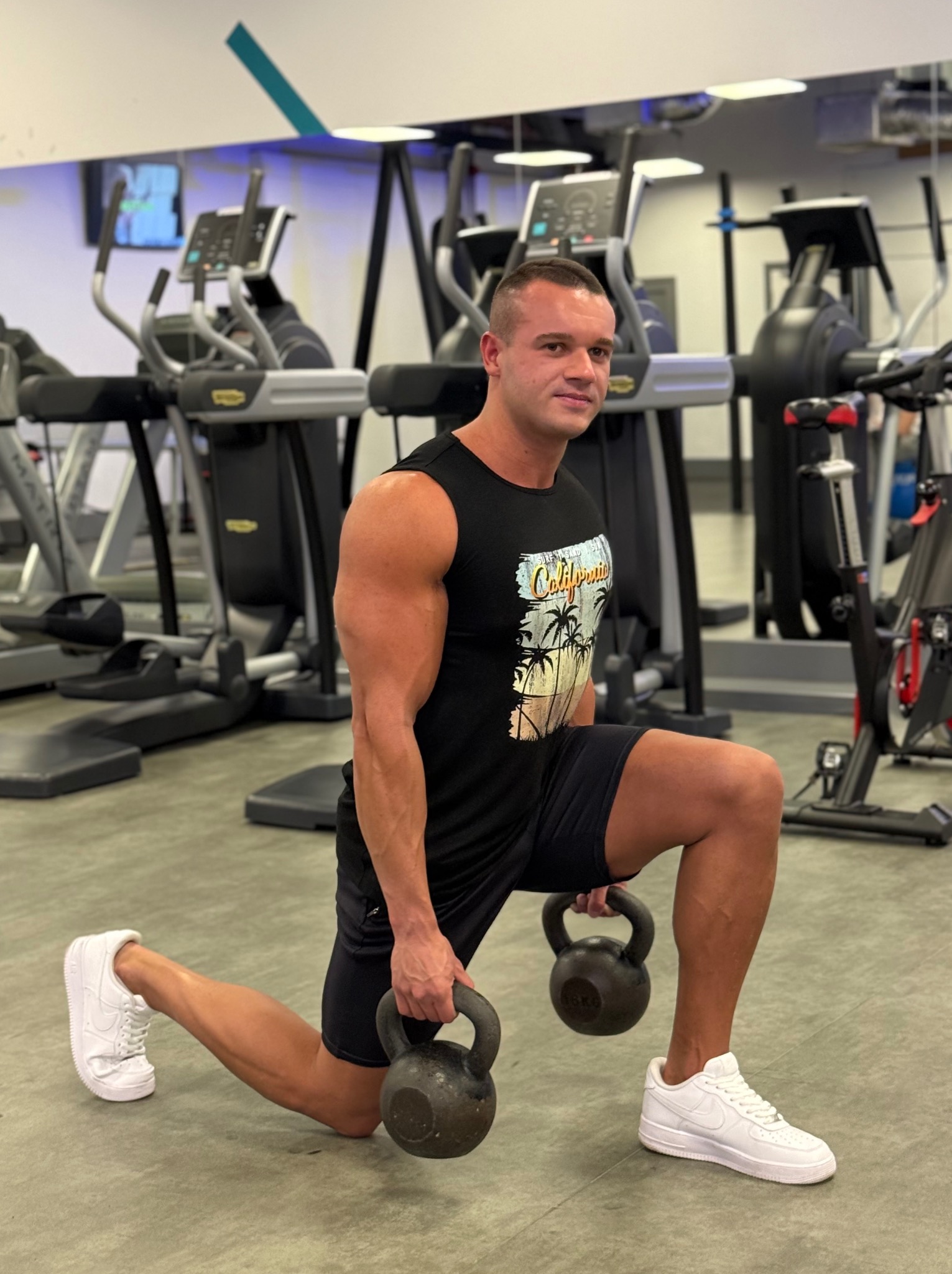 Mężczyzna w czarnej koszulce z nadrukiem California wykonuje wykrok z dwoma kettlebells w siłowni. W tle sprzęt do ćwiczeń i lustro.