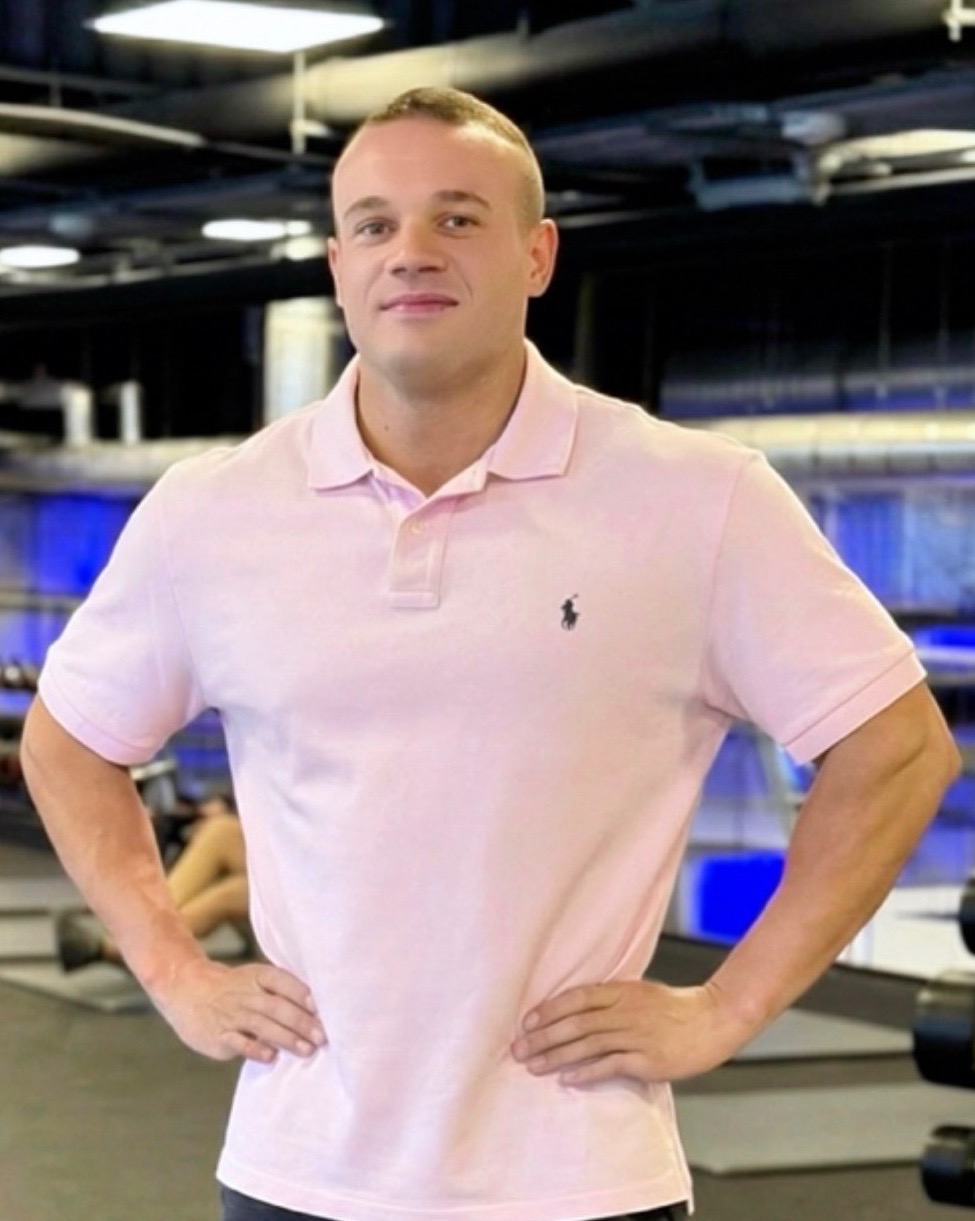 Uśmiechnięty mężczyzna w różowej koszulce polo, z rękami na biodrach, pozuje na tle siłowni z widocznymi hantlami i ćwiczącymi osobami.