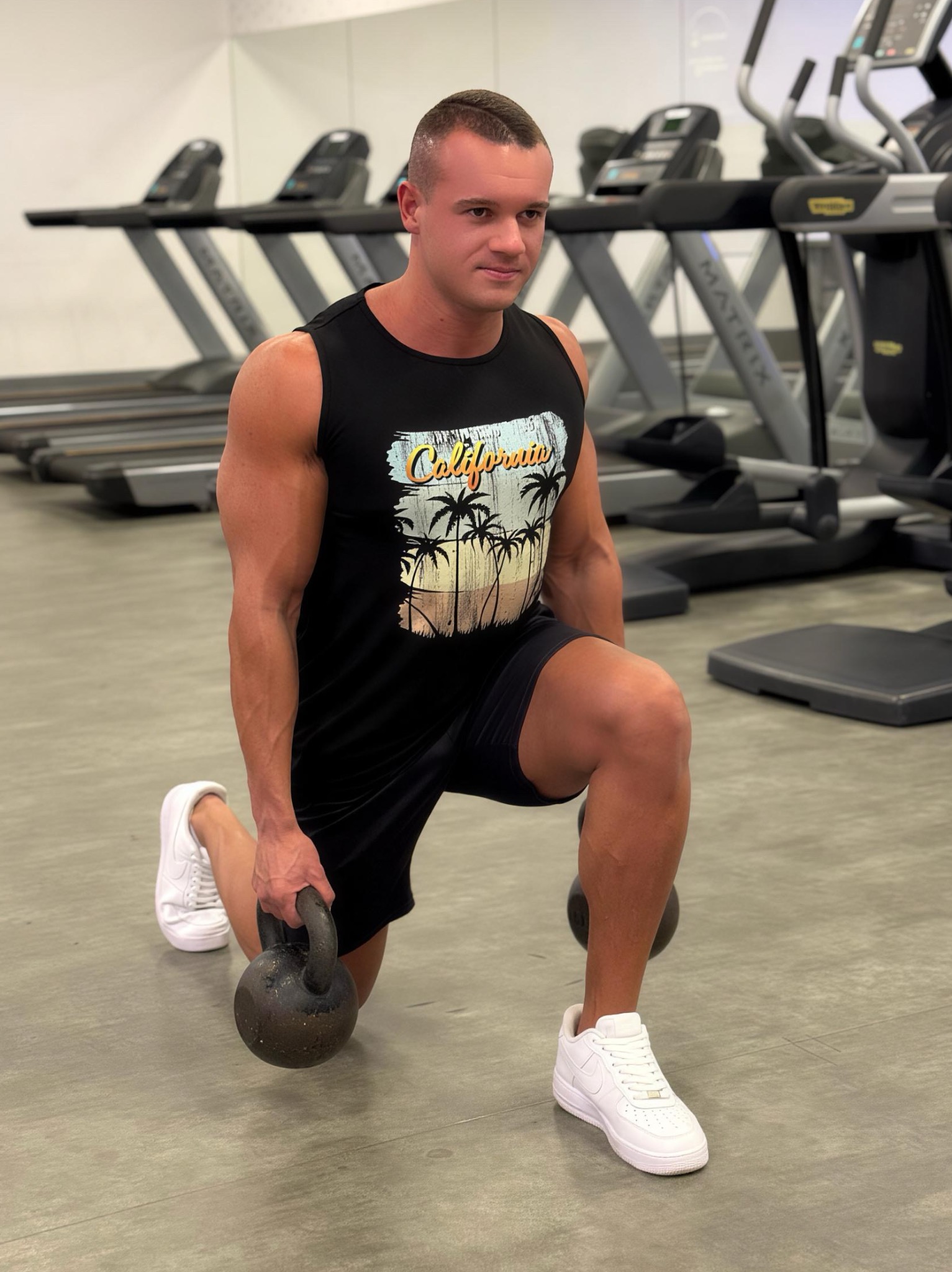 Mężczyzna w czarnej koszulce z motywem Kalifornii wykonuje wykrok z obciążeniem kettlebell w siłowni, na tle bieżnie. Białe sportowe buty.