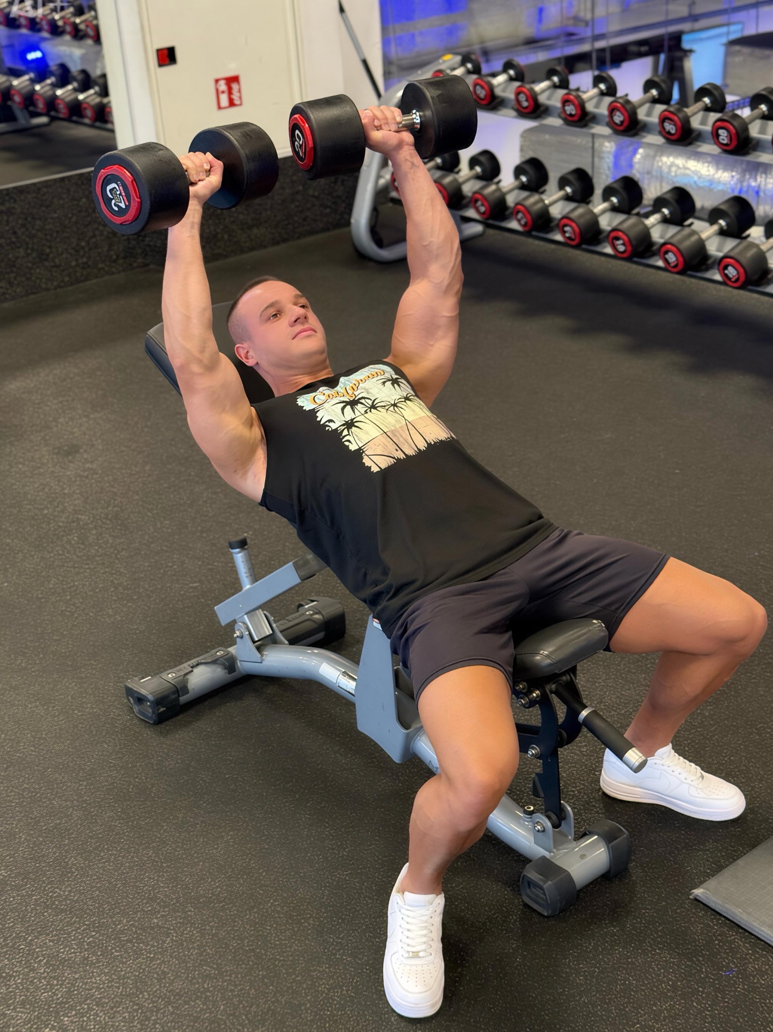 Mężczyzna na ławce w siłowni wyciska sztangielki, unosząc je nad klatką piersiową. W tle rząd hantli na stojaku i lustro.