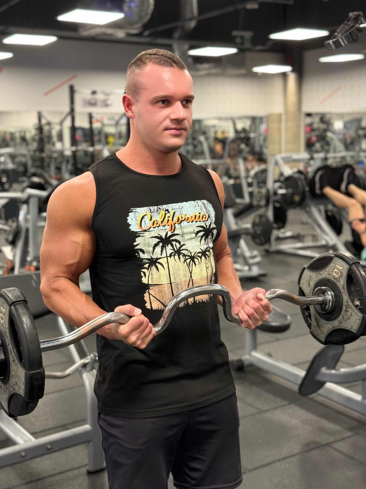 Mężczyzna w czarnej koszulce z nadrukiem California ćwiczy biceps sztangą łamaną w siłowni. Widoczne maszyny i osoby w tle, oświetlenie sufitowe.