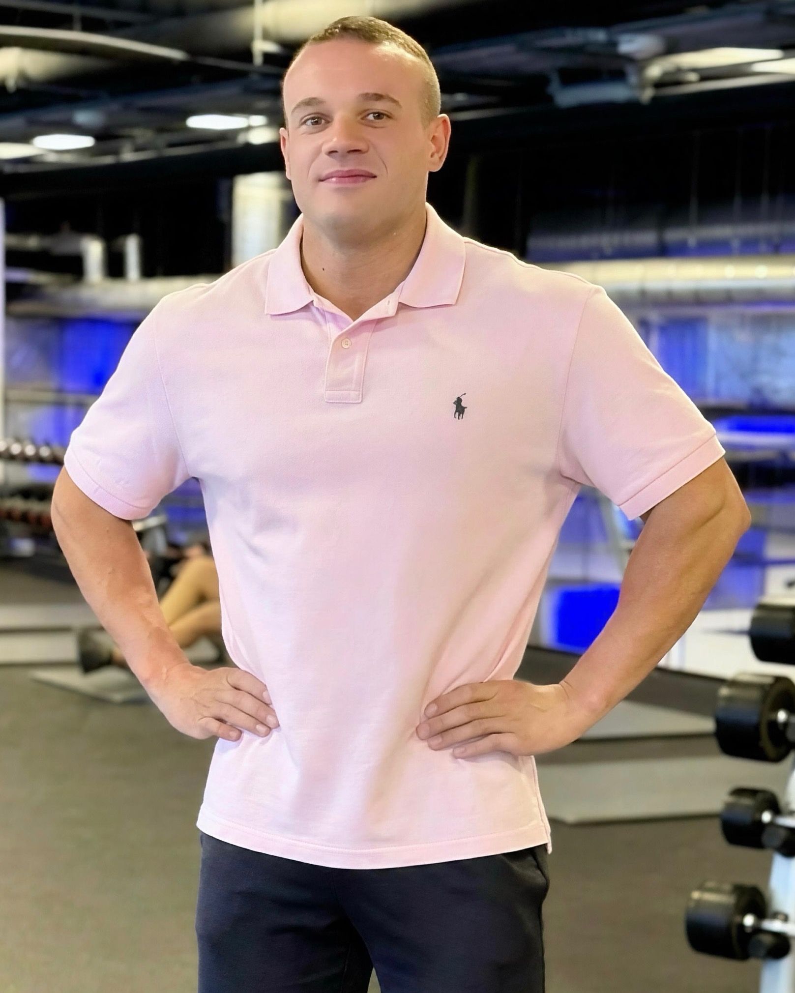 Mężczyzna w różowej koszulce polo pozuje w siłowni, ręce na biodrach. W tle widoczne sztangi i sprzęt do ćwiczeń. Dobre oświetlenie, wyraźne detale.