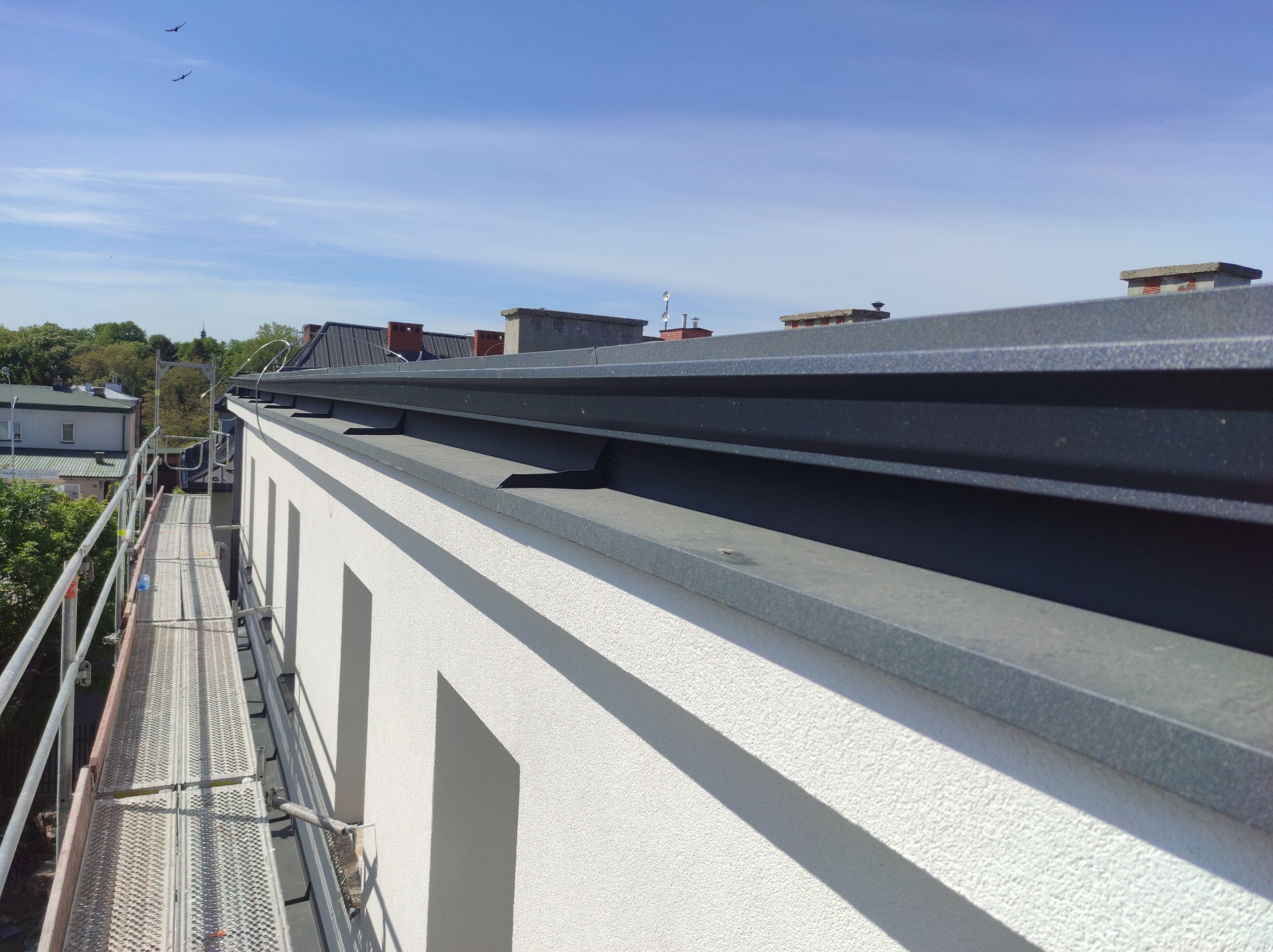 Szczegółowe ujęcie nowo zamontowanej, grafitowej obróbki blacharskiej na krawędzi dachu budynku z białą elewacją, z widocznym rusztowaniem i ptakami na niebie.