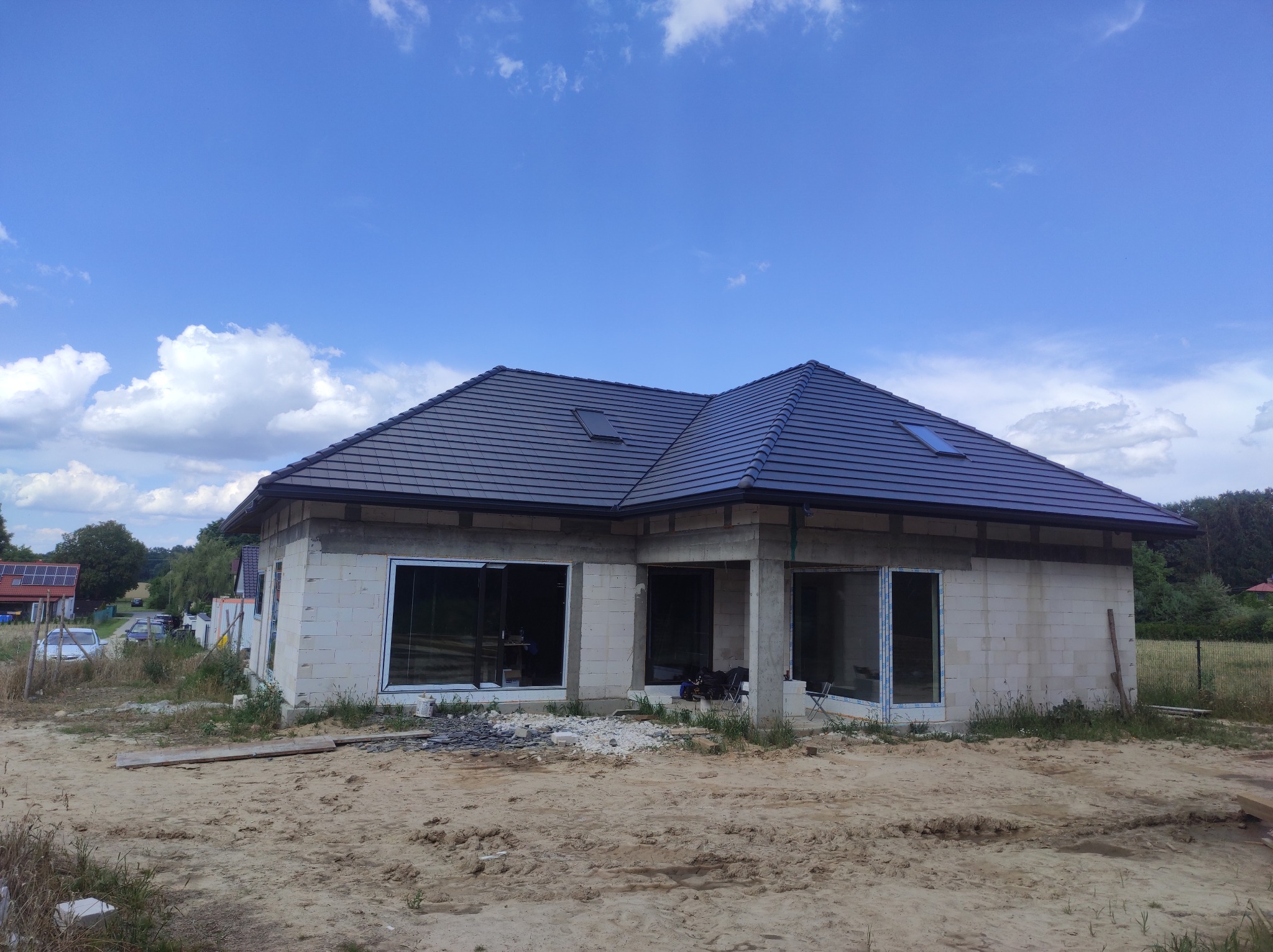 Nowoczesny dom jednorodzinny w trakcie budowy, widoczny dach z ciemnej dachówki ceramicznej z dwoma oknami dachowymi, ściany z białego betonu komórkowego, duże okna wstawione, teren wokół domu...