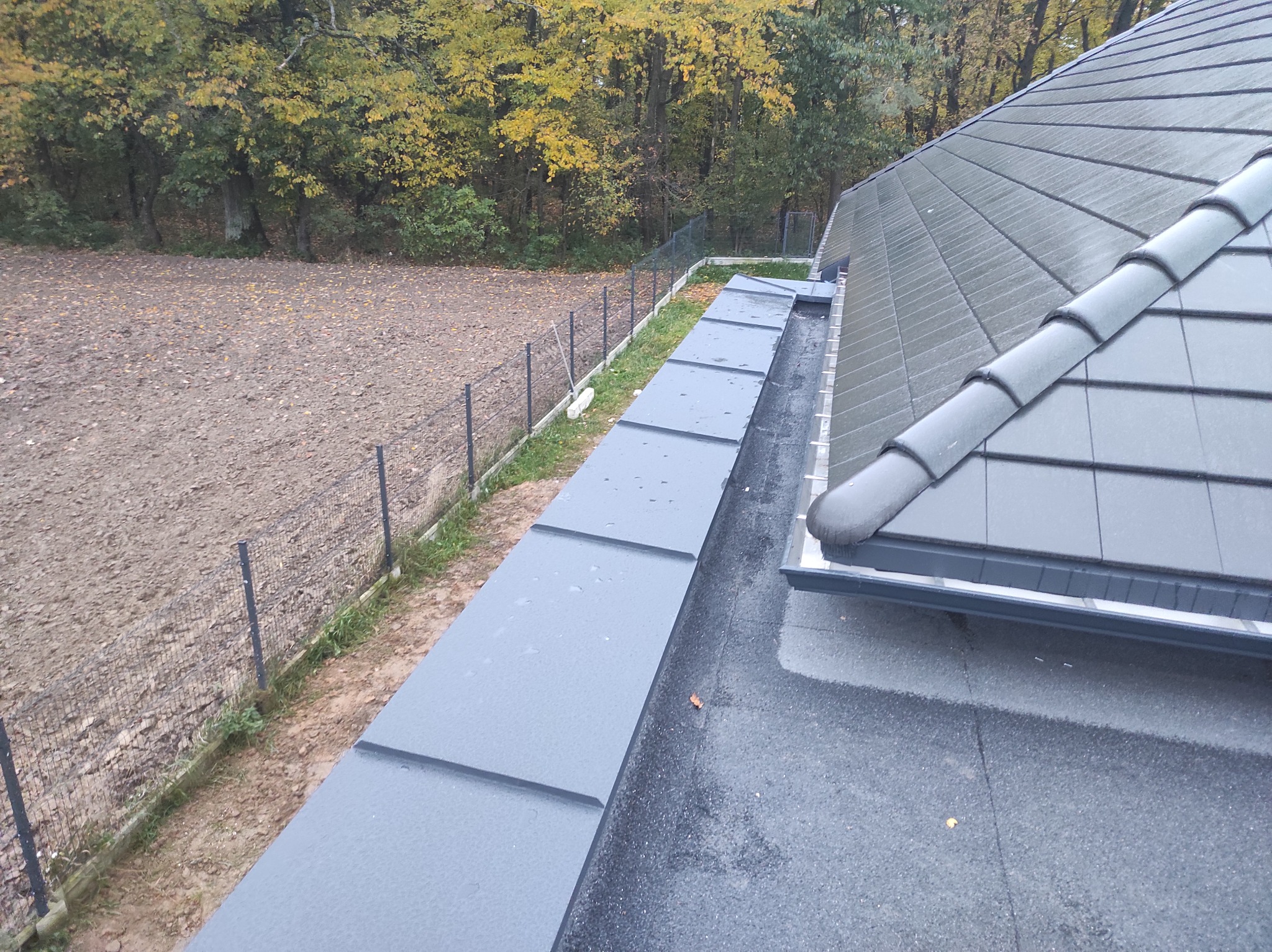Nowoczesny dach z szarej dachówki ceramicznej, widoczny fragment obróbki blacharskiej i rynny, tło stanowią jesienne drzewa i pole.