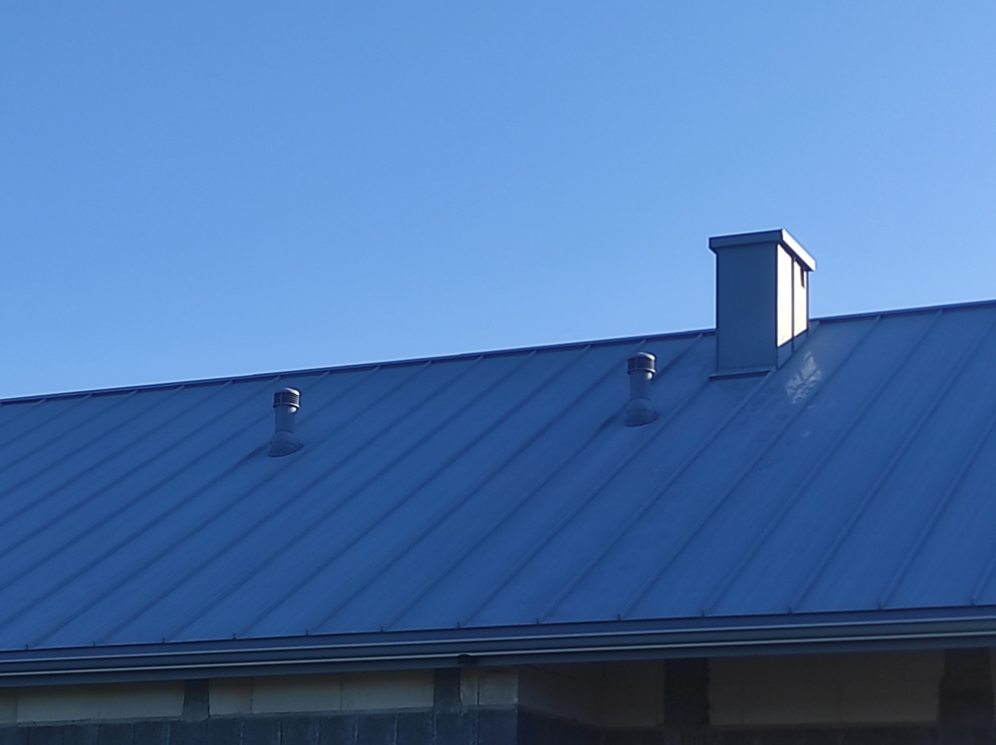 Nowy, metalowy dach w kolorze grafitowym z dwoma odpowietrznikami i kominem na tle błękitnego nieba.