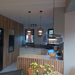 StyleHouse - Nowoczesna kuchnia z wyspą, oświetlona wiszącymi lampami o dymionym szkle, widok na schody i ogród przez okno, drewniane wykończenie szafek.